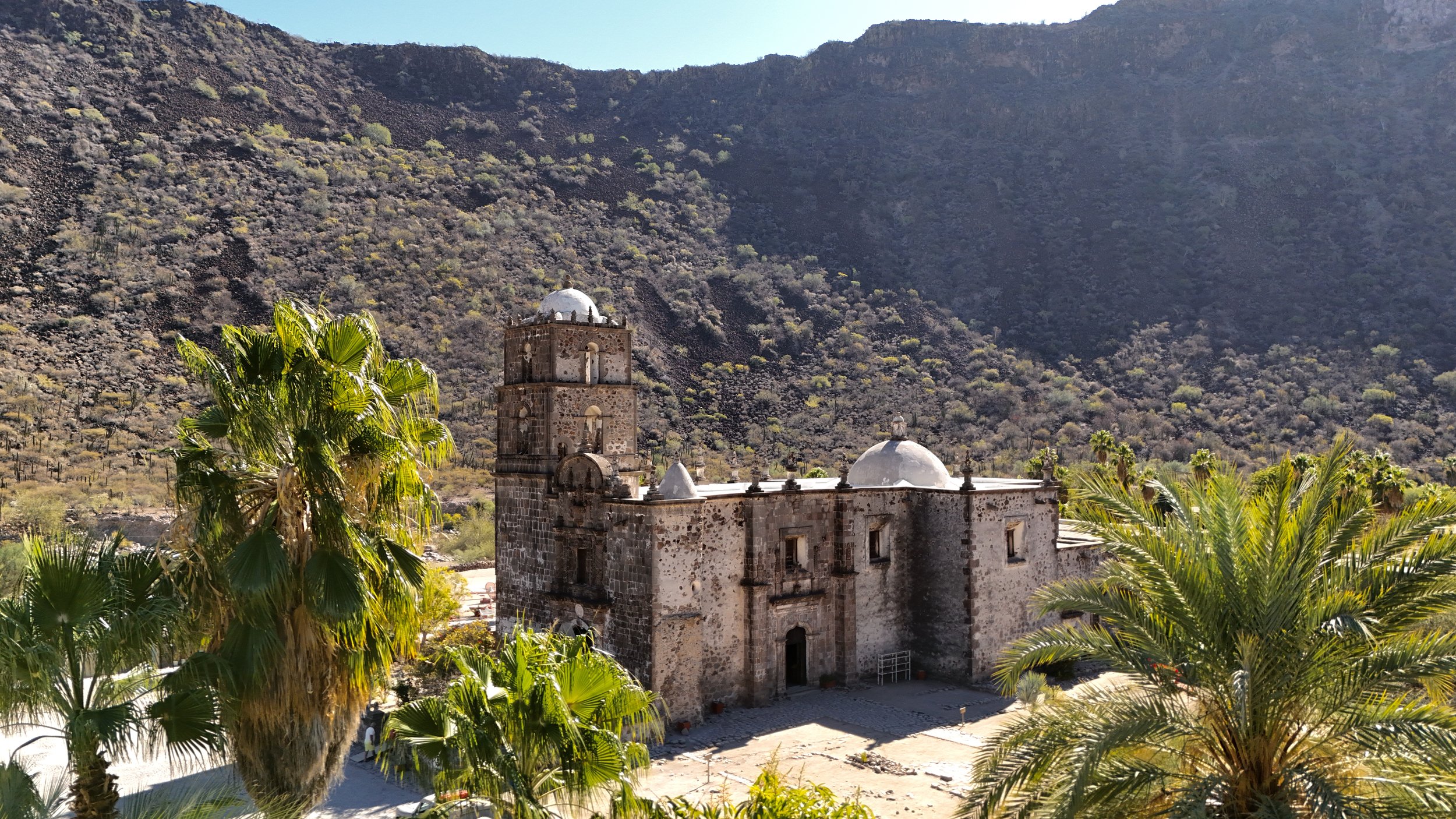 Historic stone church with domed roofs, surrounded by palm trees, set against arid mountains in the background during daylight.