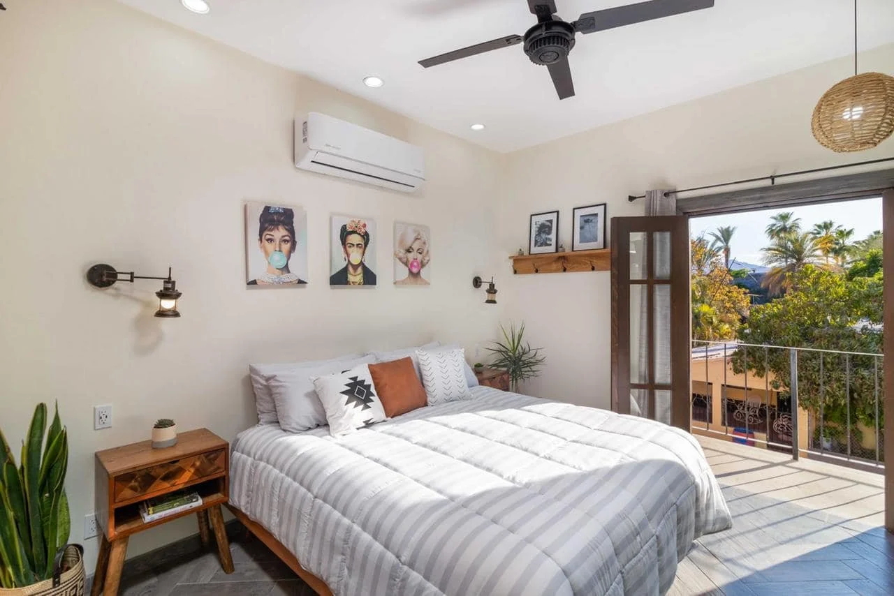 Bright bedroom with a white bed, wooden nightstand, artwork on the wall, two wall-mounted lamps, a ceiling fan, an air conditioning unit, a plant, and an open balcony door revealing outdoor trees and palm trees.