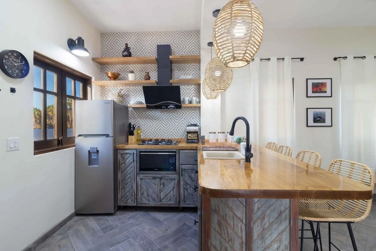 Kitchen with wooden accents, white walls, stainless steel refrigerator, and hanging wicker lamps. Open shelves with decorative items, patterned backsplash, and bar seating area with four wicker chairs.