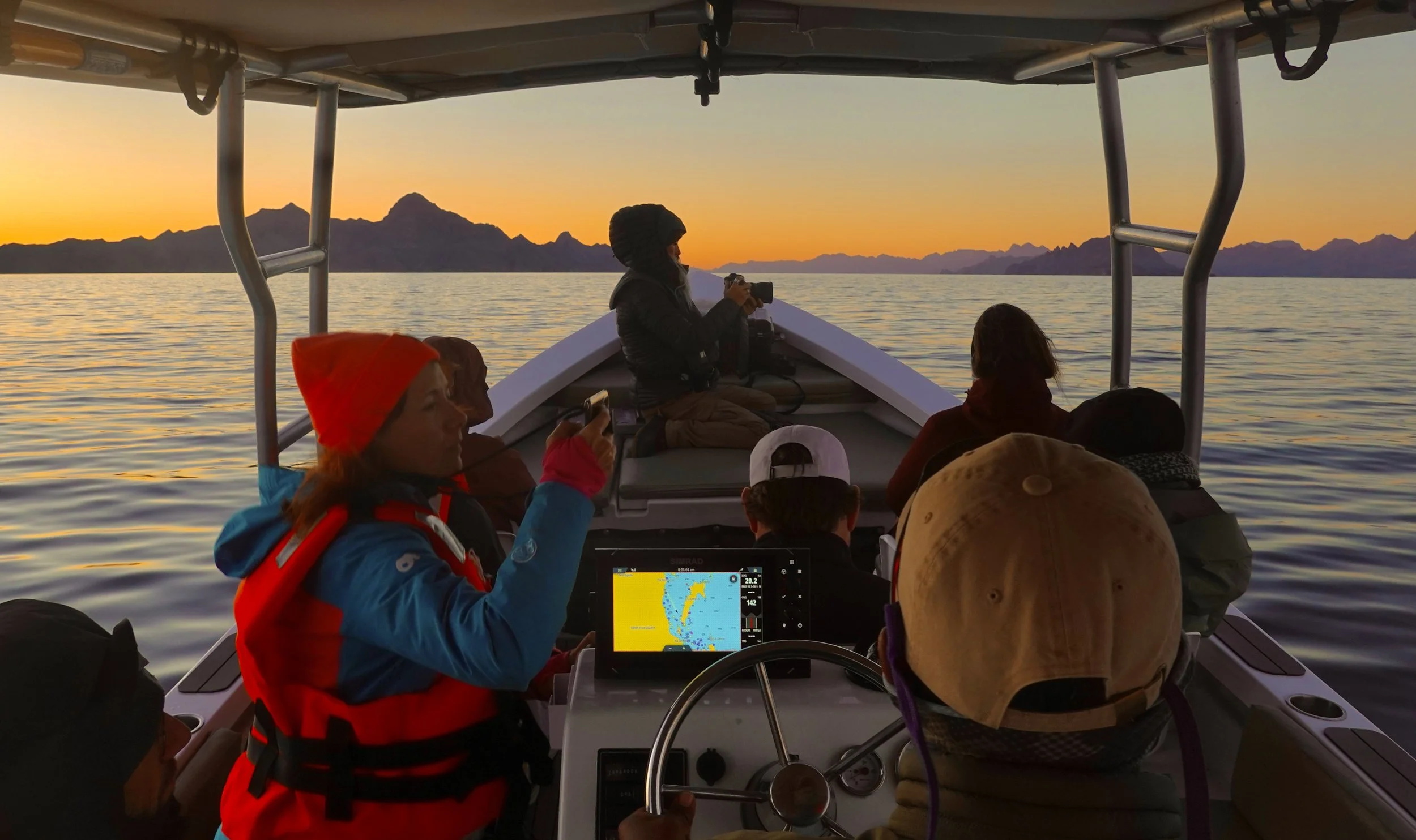 A group of people on a boat during sunset, with a mountain range in the background. Some are taking photos or using devices, and there is a fish finder screen showing underwater data.