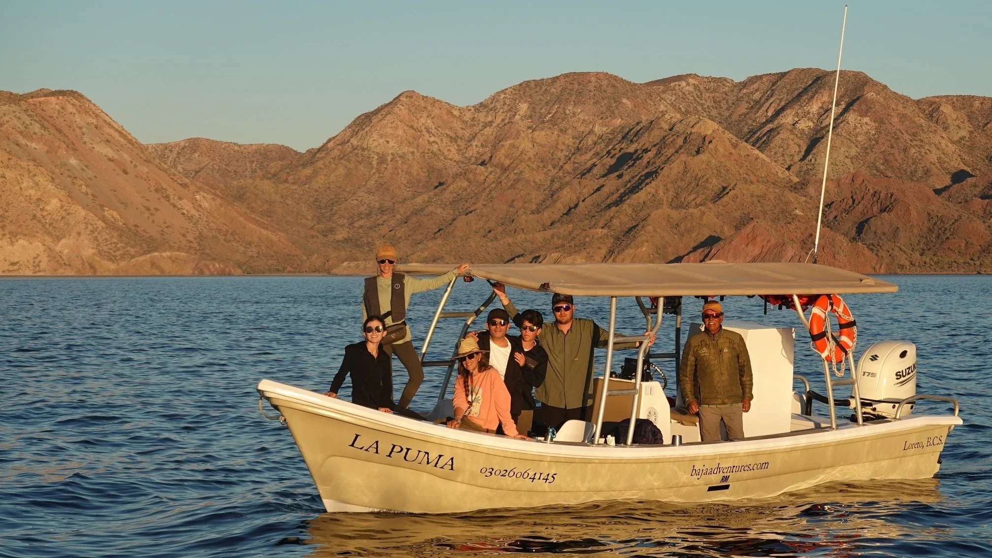A group of eight people on a boat named La Puma, floating on a lake with mountainous terrain in the background, during sunset.