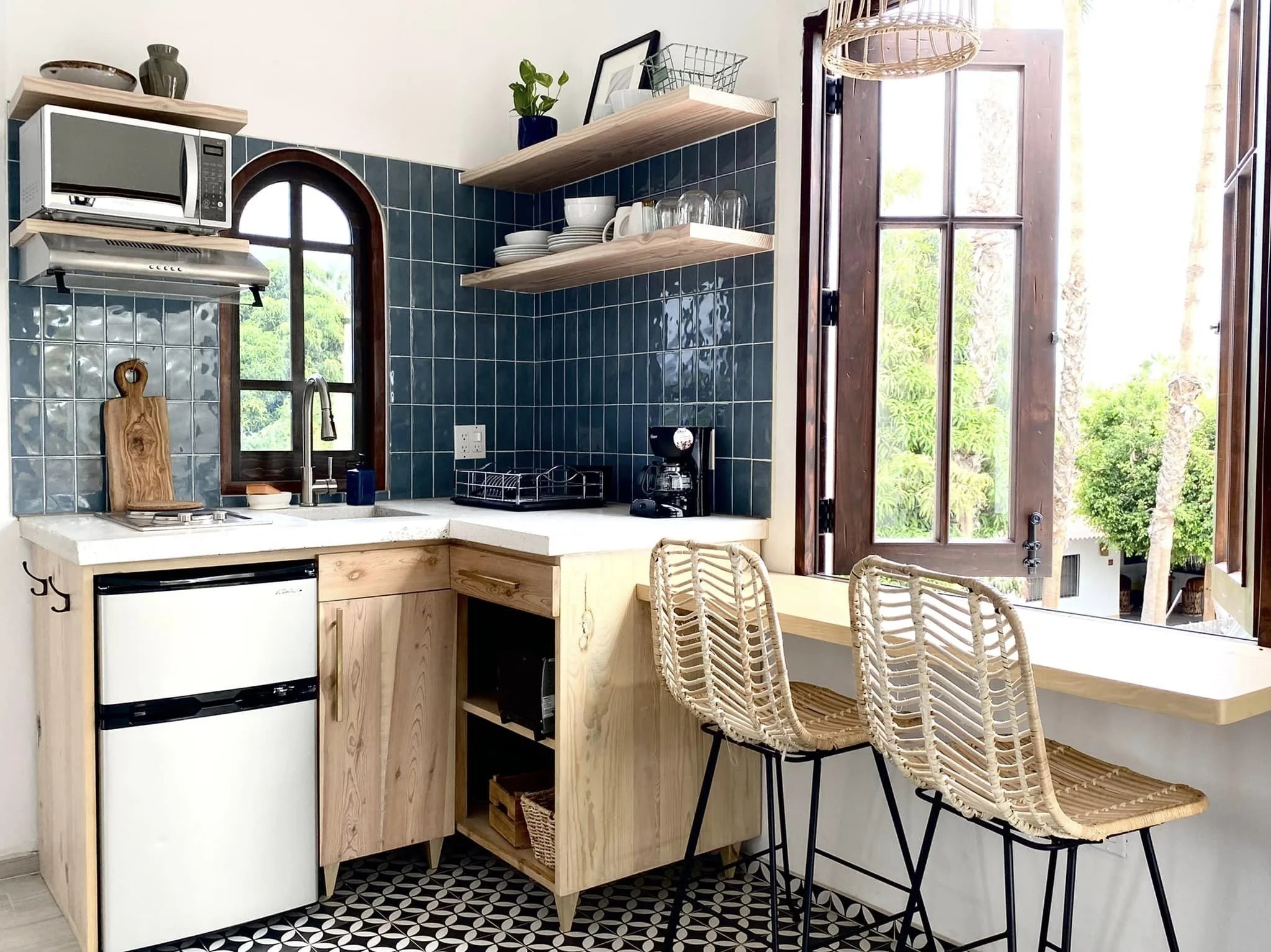 Kitchen with a small fridge, mosaic tile counter, open shelves with dishes, coffee maker, microwave, open window showing outdoor greenery, and a bar with three rattan chairs.