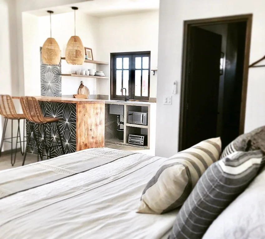 View of a cozy living space with a bed in the foreground, a kitchen area with bar stools, open shelves, and a small window in the background.