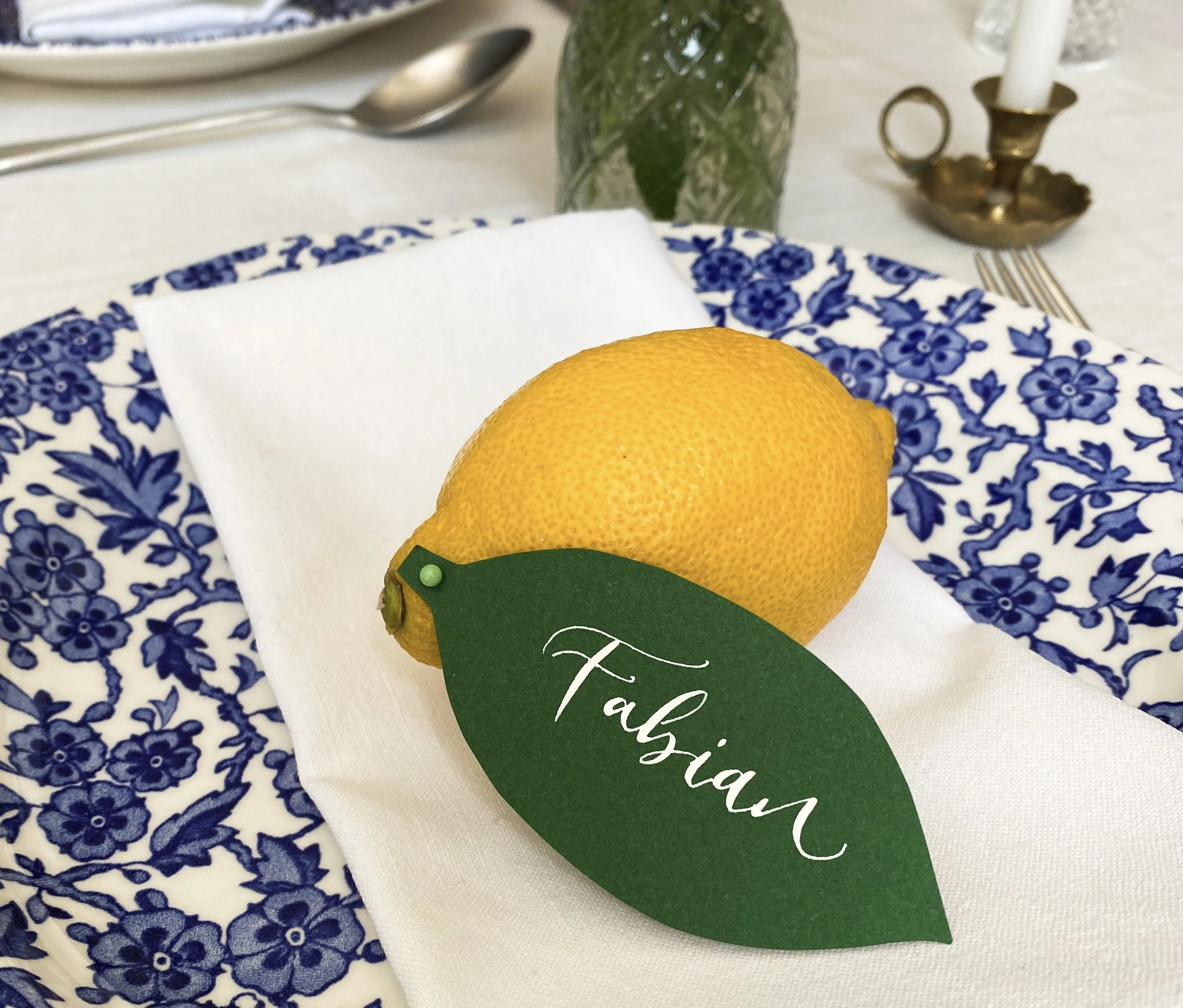 White calligraphy on a green leaf, pinned to a lemon. The calligraphy is a relaxed, modern style and the lemon and leaf is arrange on a napkin, on a plate on a table set for eating.
