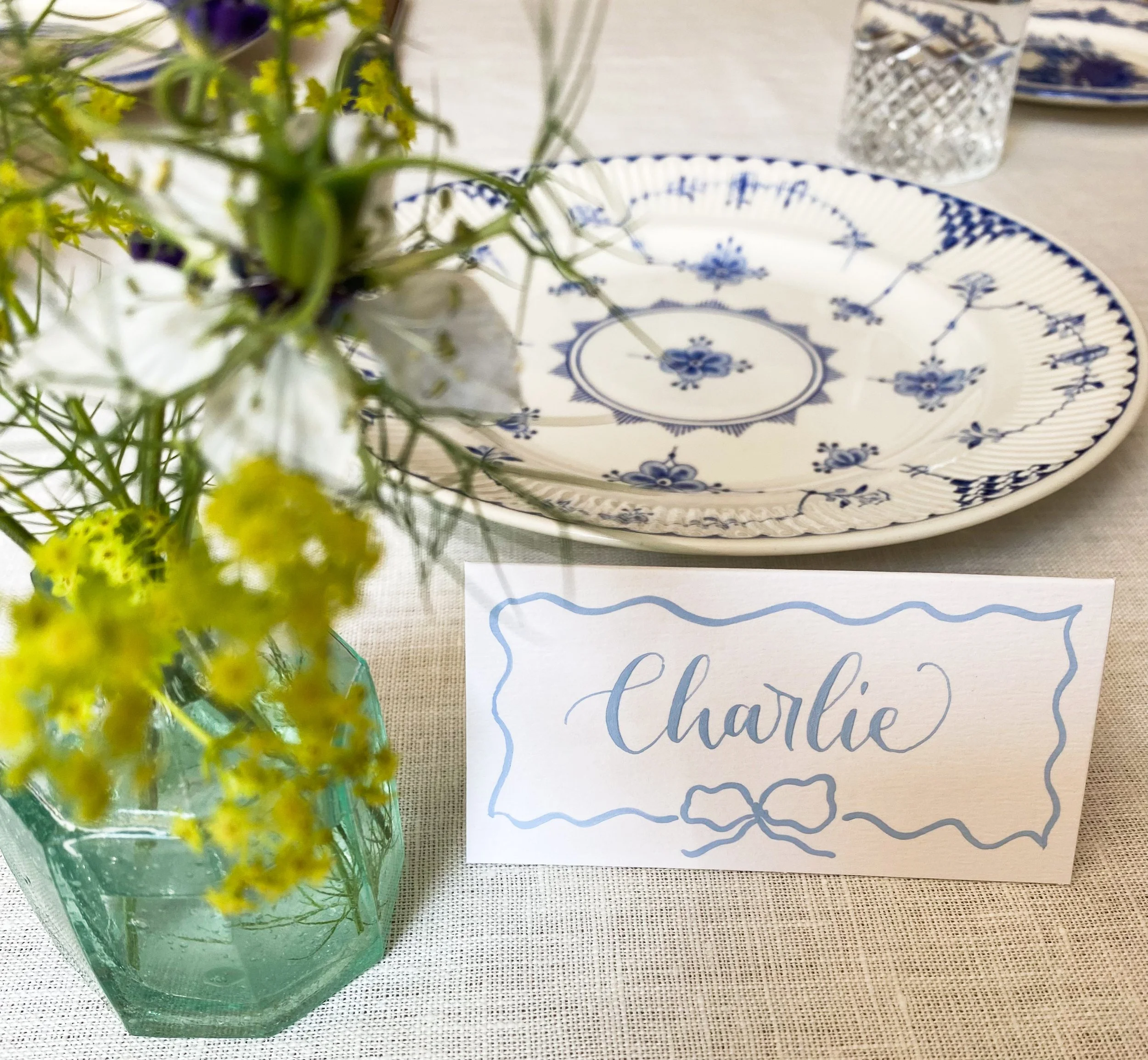 More traditional blue calligraphy on a white placecard with a painted wavy bow design. The place card is styled on a table with flowers and a plate.