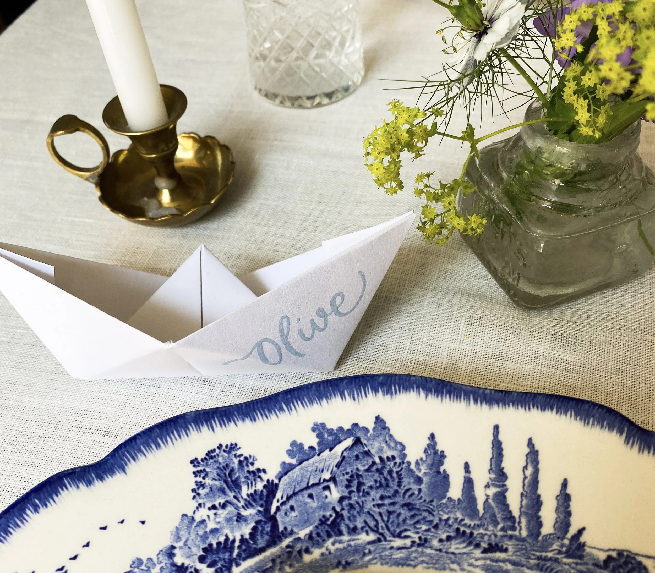 Blue brush lettering calligraphy on the side of an origami boat styled on a table set with brass chamber stick and flowers.