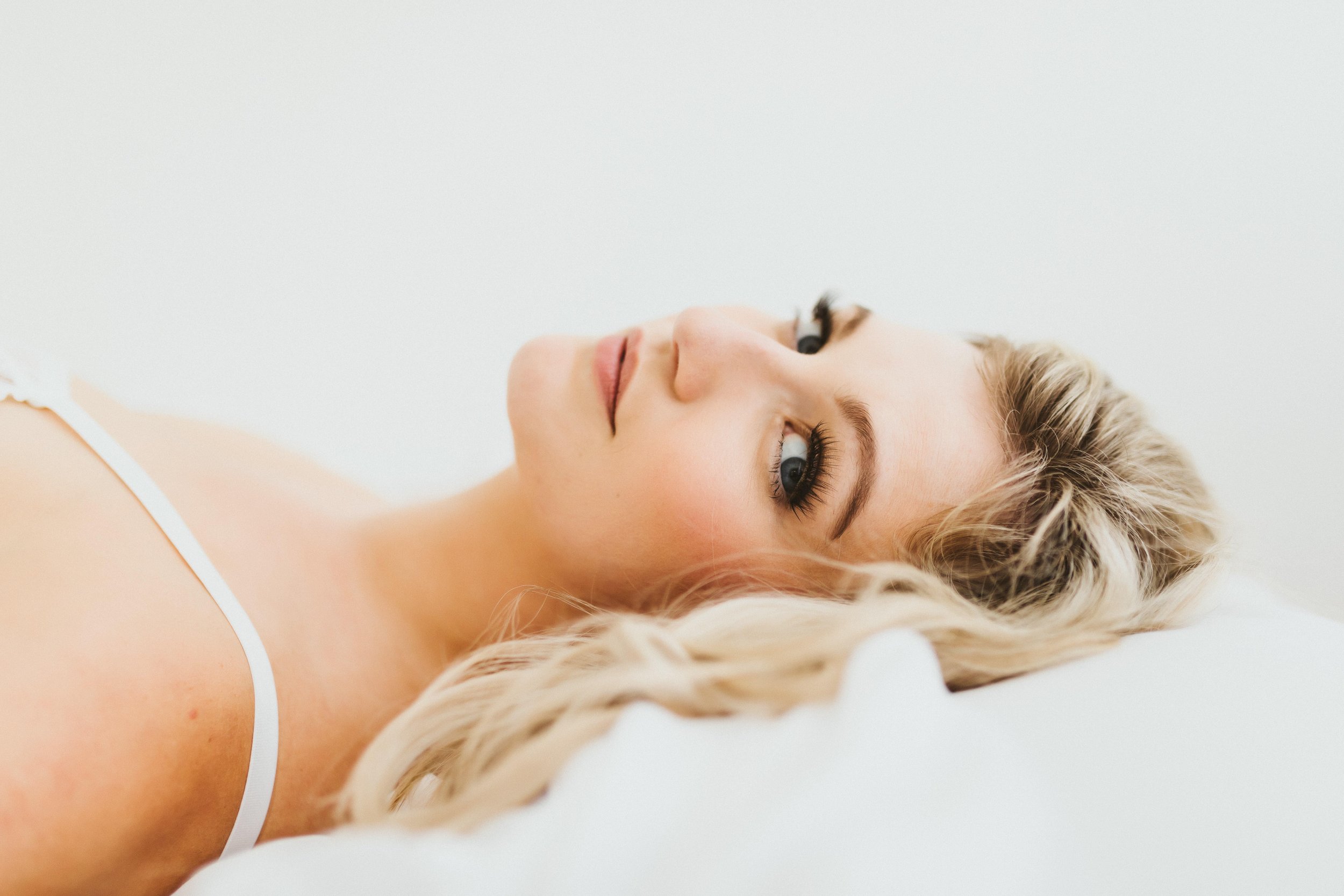 A wedding bridal fiancee during a lingerie photo shoot laying on white sheets. 