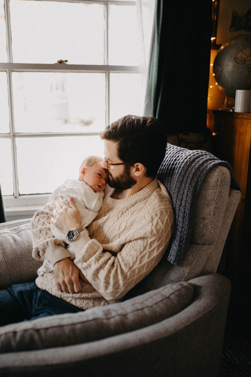 Dad relaxing with newborn son in lounge chair near a window for lifestyle photos. 