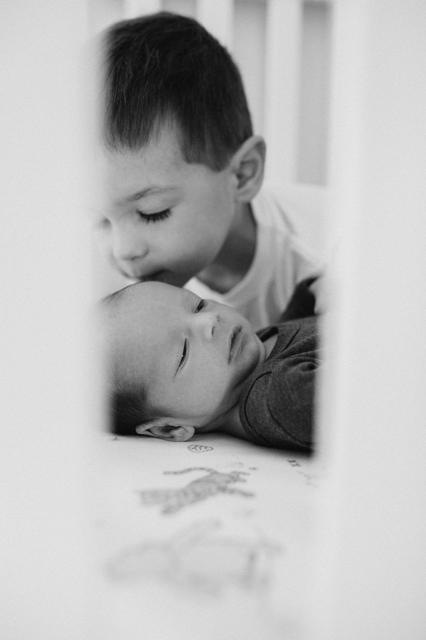 Big brother kissing baby on the head in the crib of Lakewood home during photo session.