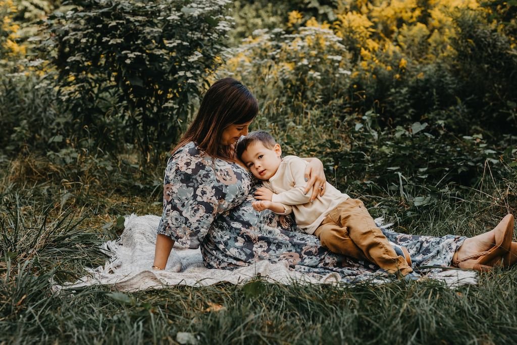 Pregnant mother laying on grass with toddler son posing for outdoor maternity pictures. 