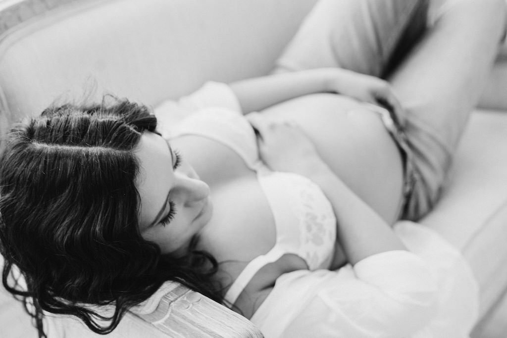 Pregnant woman laying on sofa with unbuttoned shirt showing her belly during a maternity shoot.