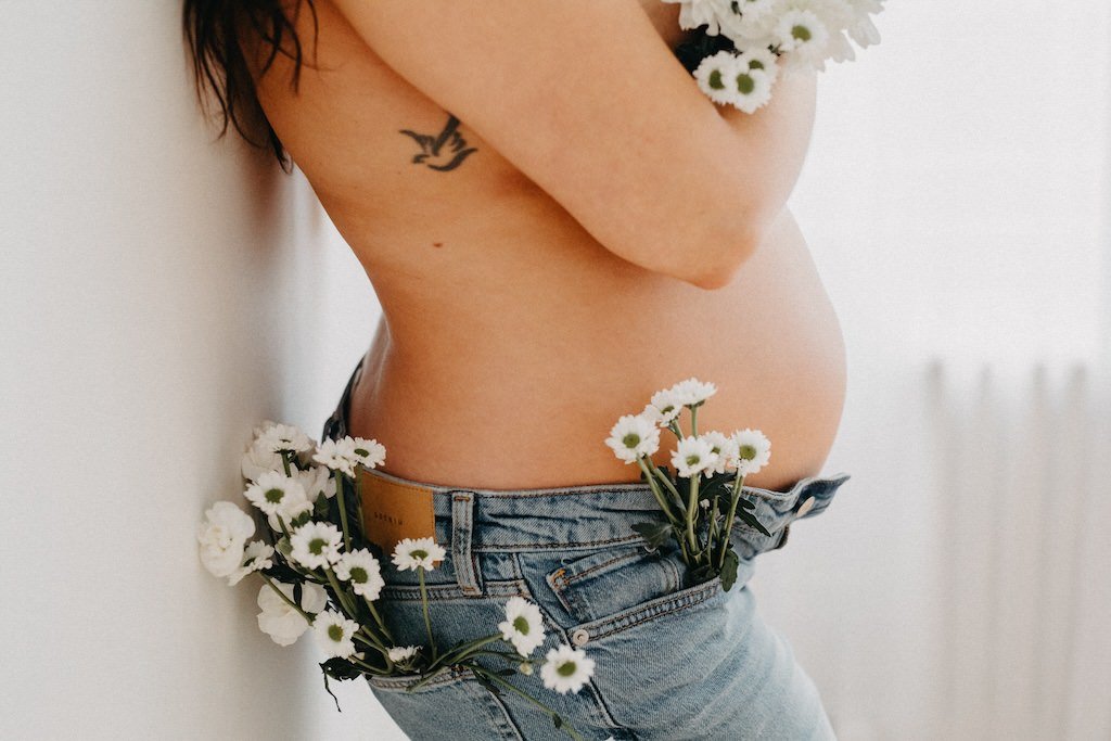 Pregnant woman wearing jeans and decorated with flower over her breasts and waistline during a studio maternity photo shoot. 