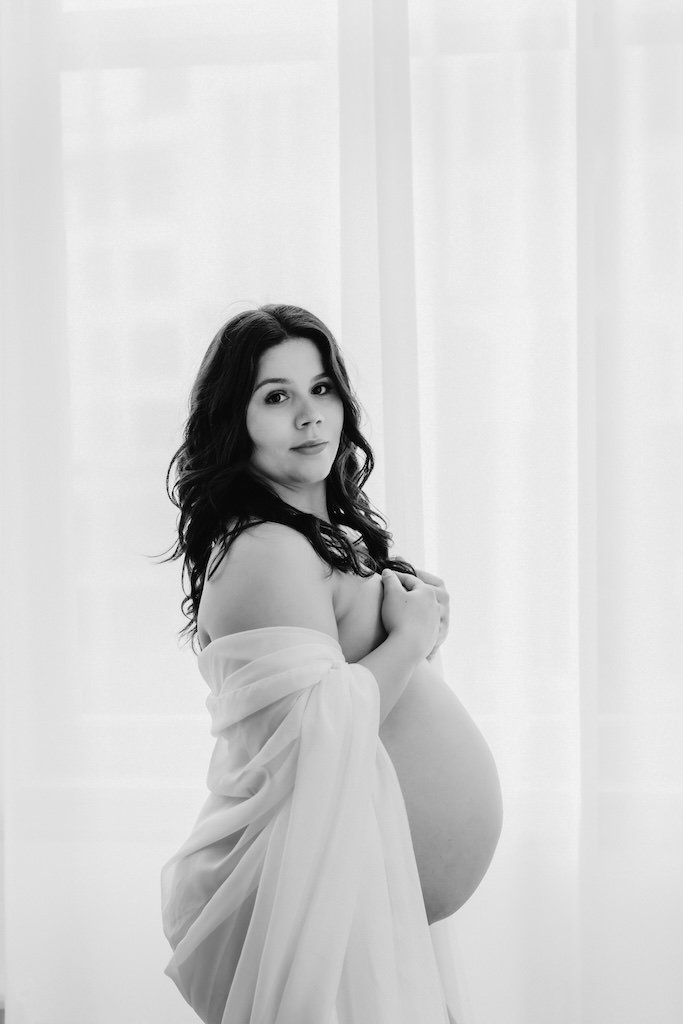 Studio black and white maternity shoot of woman in black hair wearing white chiffon fabric. 