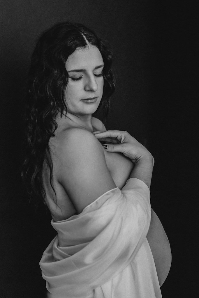 Modern photograph of pregnant woman posing by black backdrop in nude maternity shoot. 