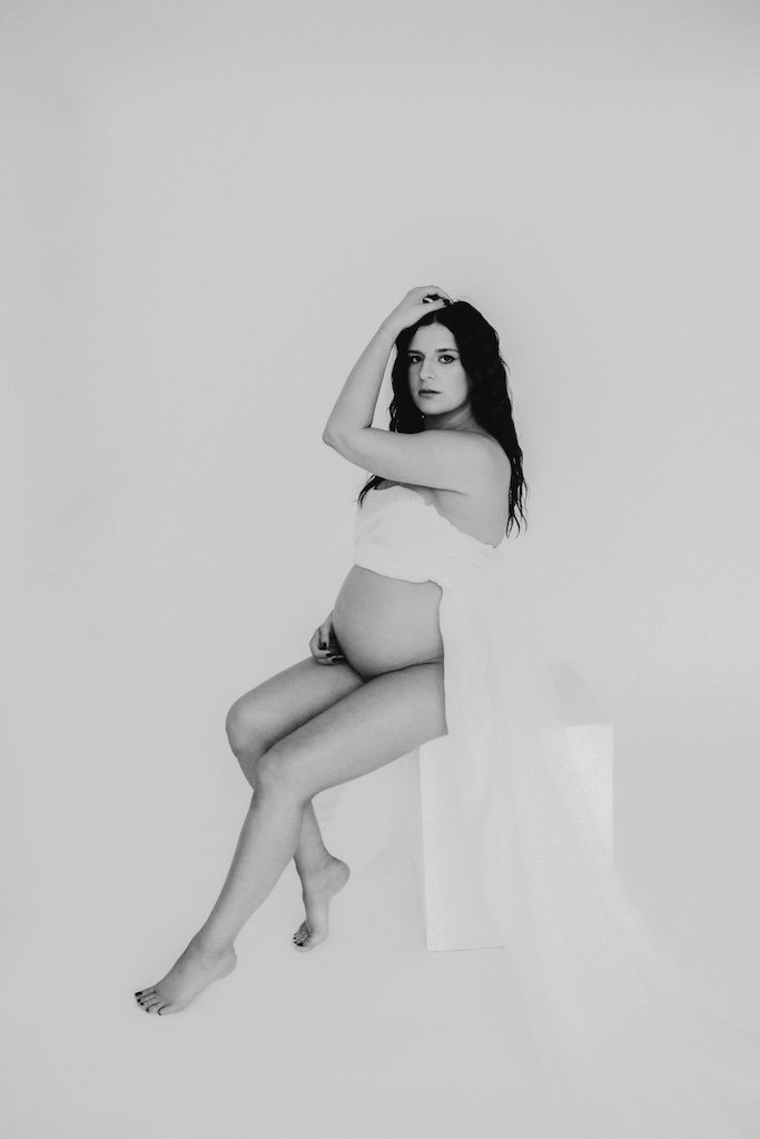 Studio maternity portrait in black and white of woman sitting on a posing cube. 