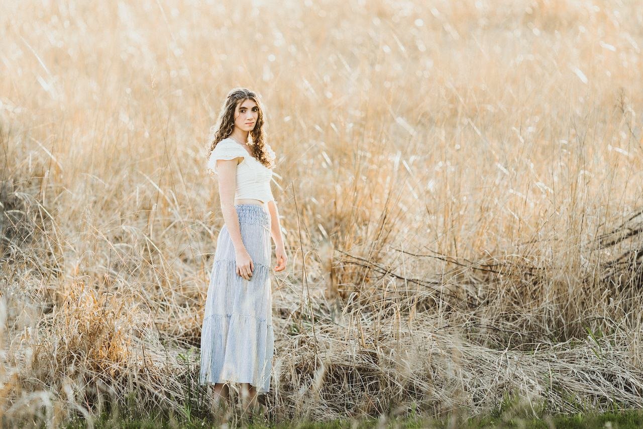 A girl wearing long skirt posing amongst tall yellow dress on a sunny day for senior pictures. 