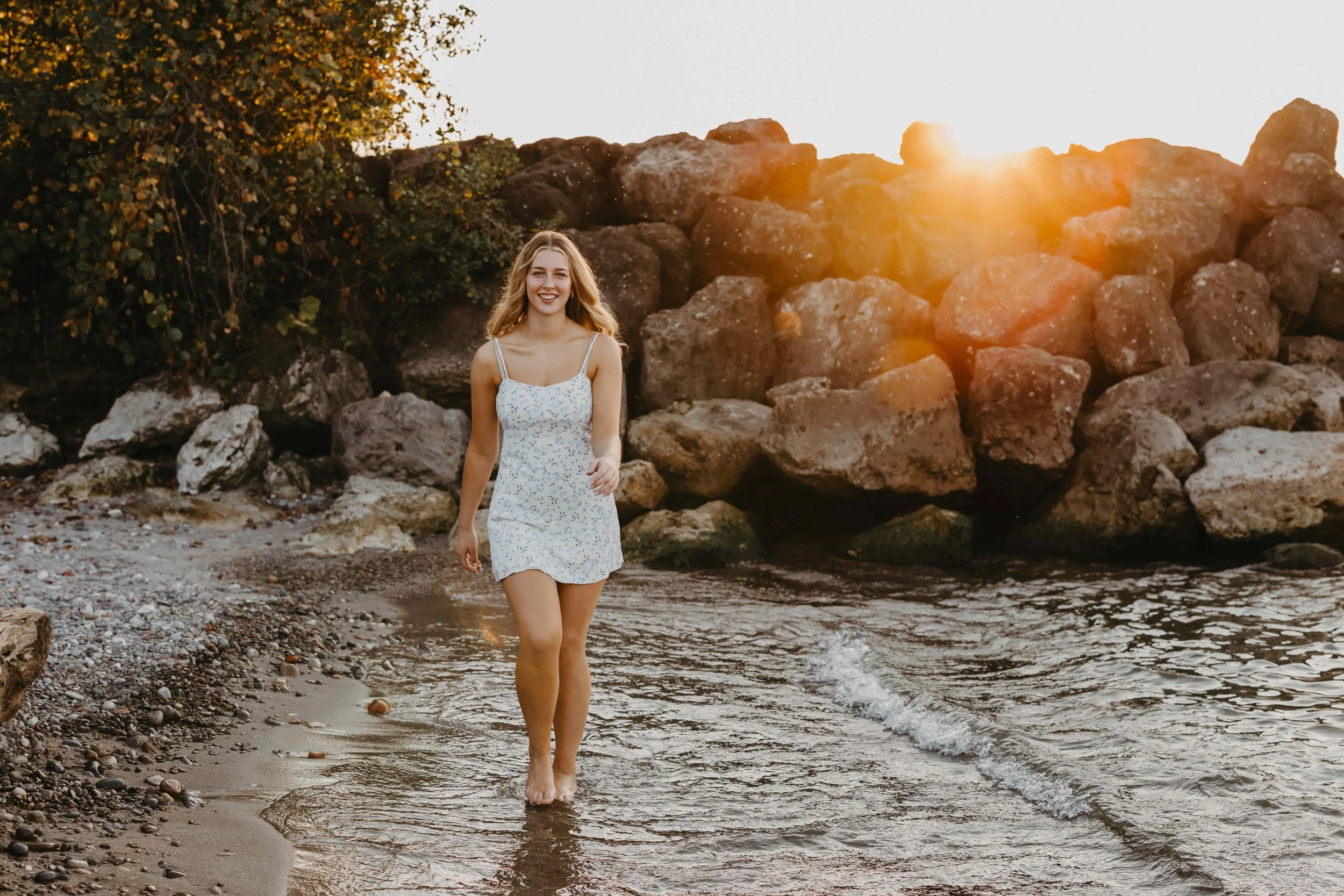 High school senior girl walking in the water at a Cleveland beach during sunset, wearing a white floral dress with large rocks in the background.