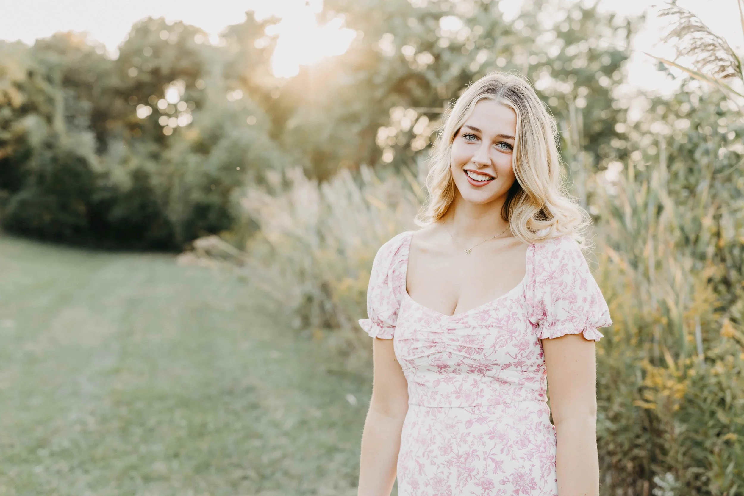 Smiling high school senior girl in a pink and white floral dress standing in a grassy field in Cleveland during golden hour.