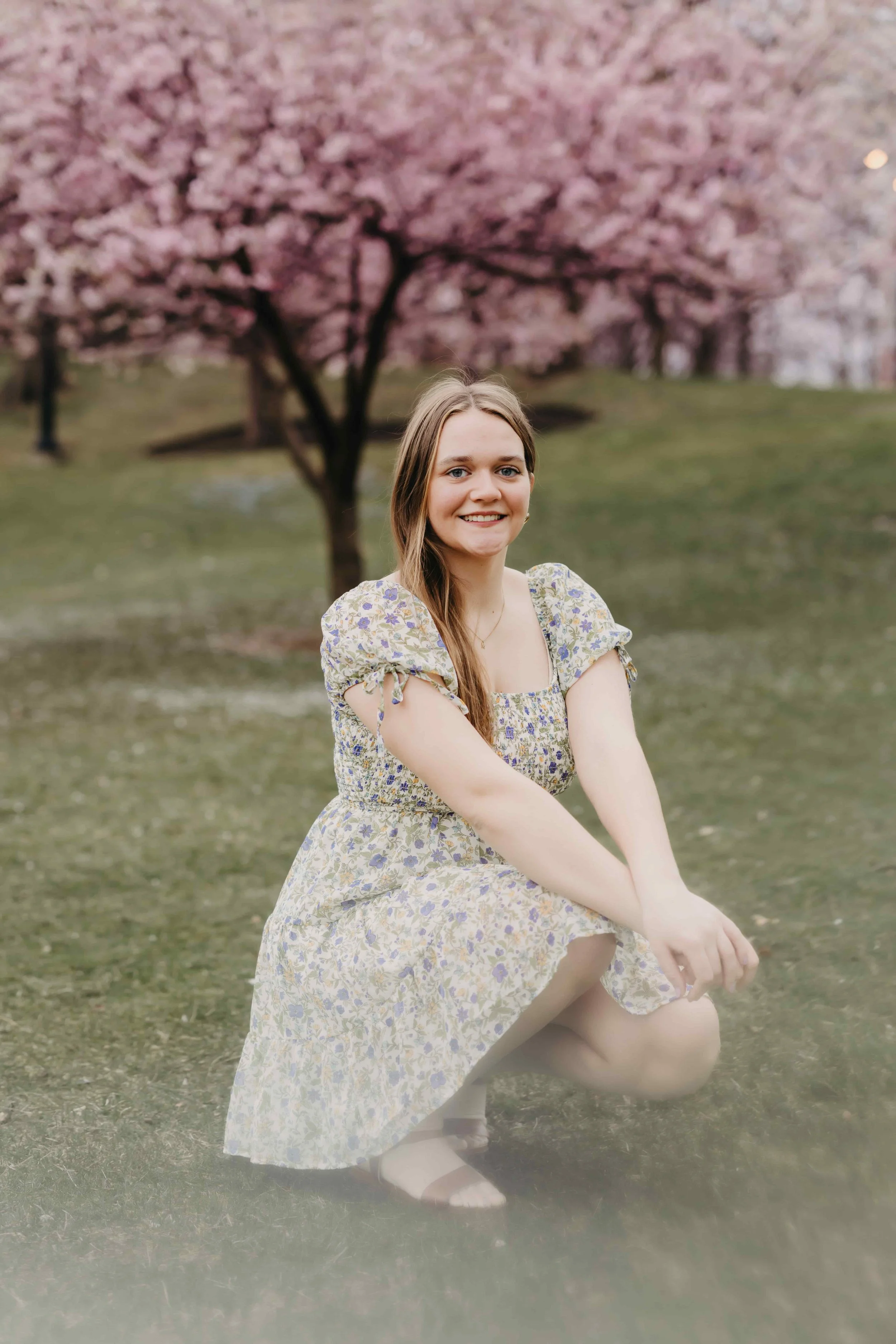 High school senior girl crouching and smiling in a park with pink cherry blossom trees in Cleveland, wearing a floral spring dress.