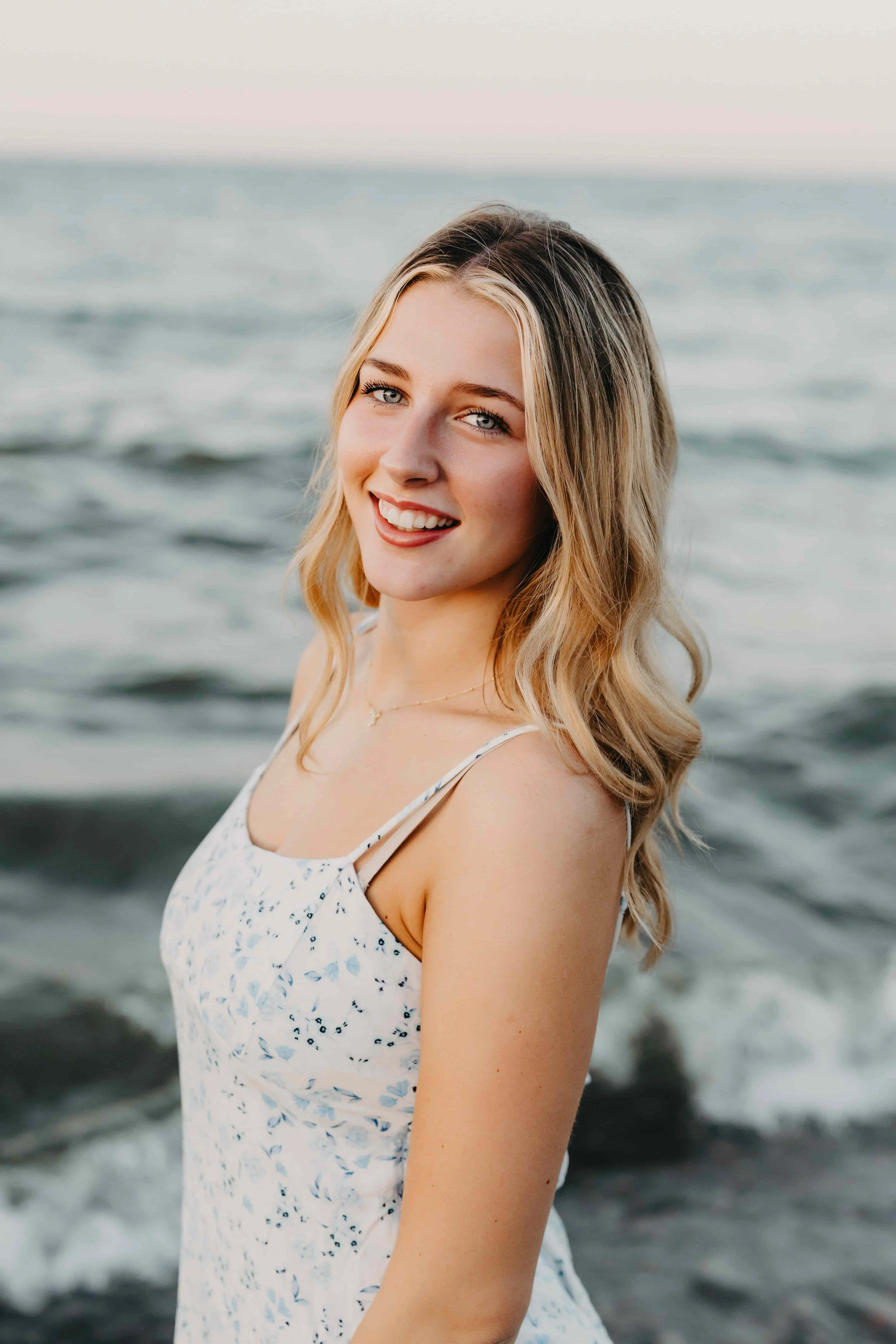 High school senior girl smiling over her shoulder at a Cleveland beach with Lake Erie waves in the background, wearing a white floral sundress.
