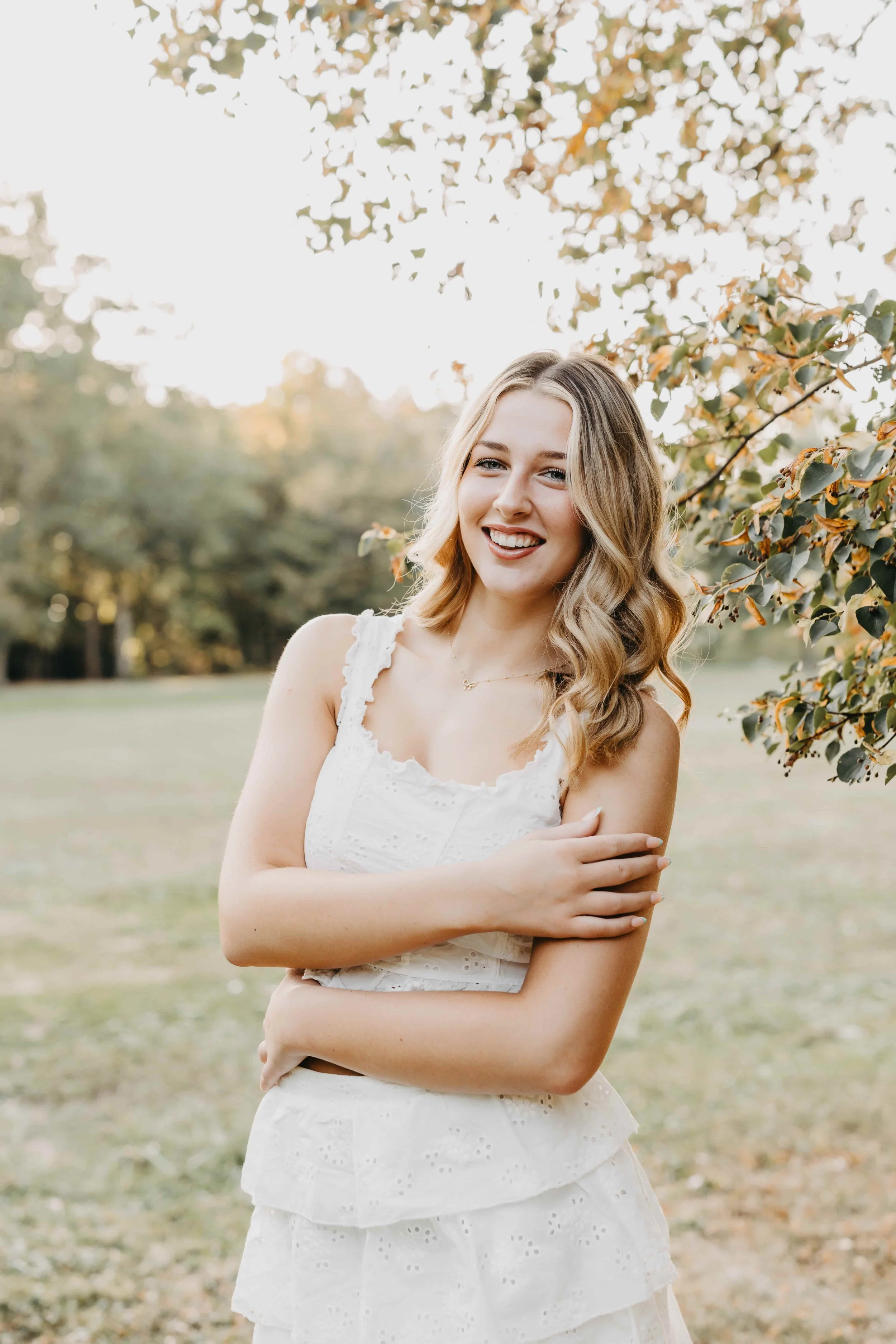 High school senior girl smiling in a white eyelet outfit standing under a tree in a Cleveland park during golden hour.