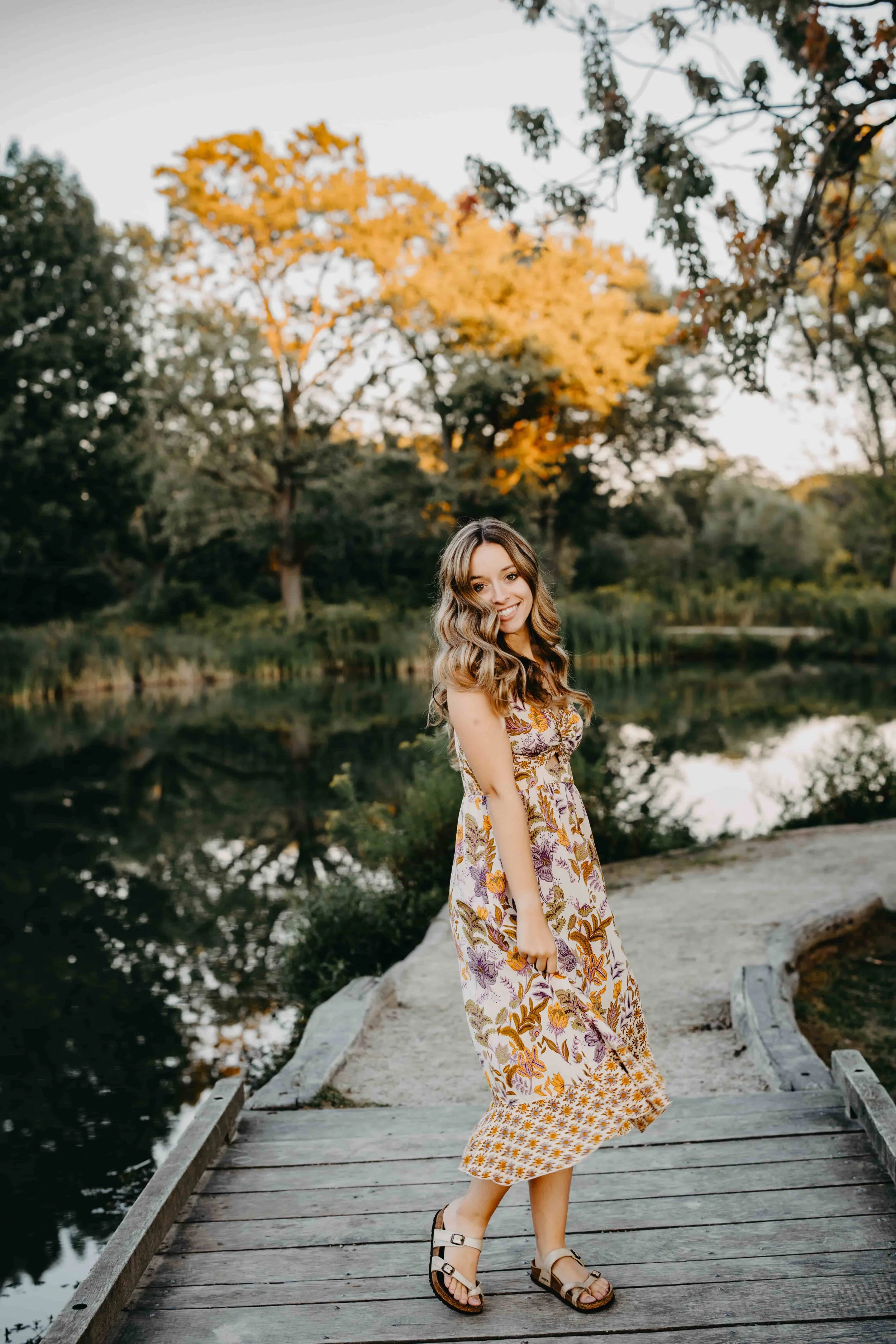 High school senior girl smiling on a wooden boardwalk over a pond with fall foliage in Cleveland, Ohio, wearing a floral midi dress and sandals.