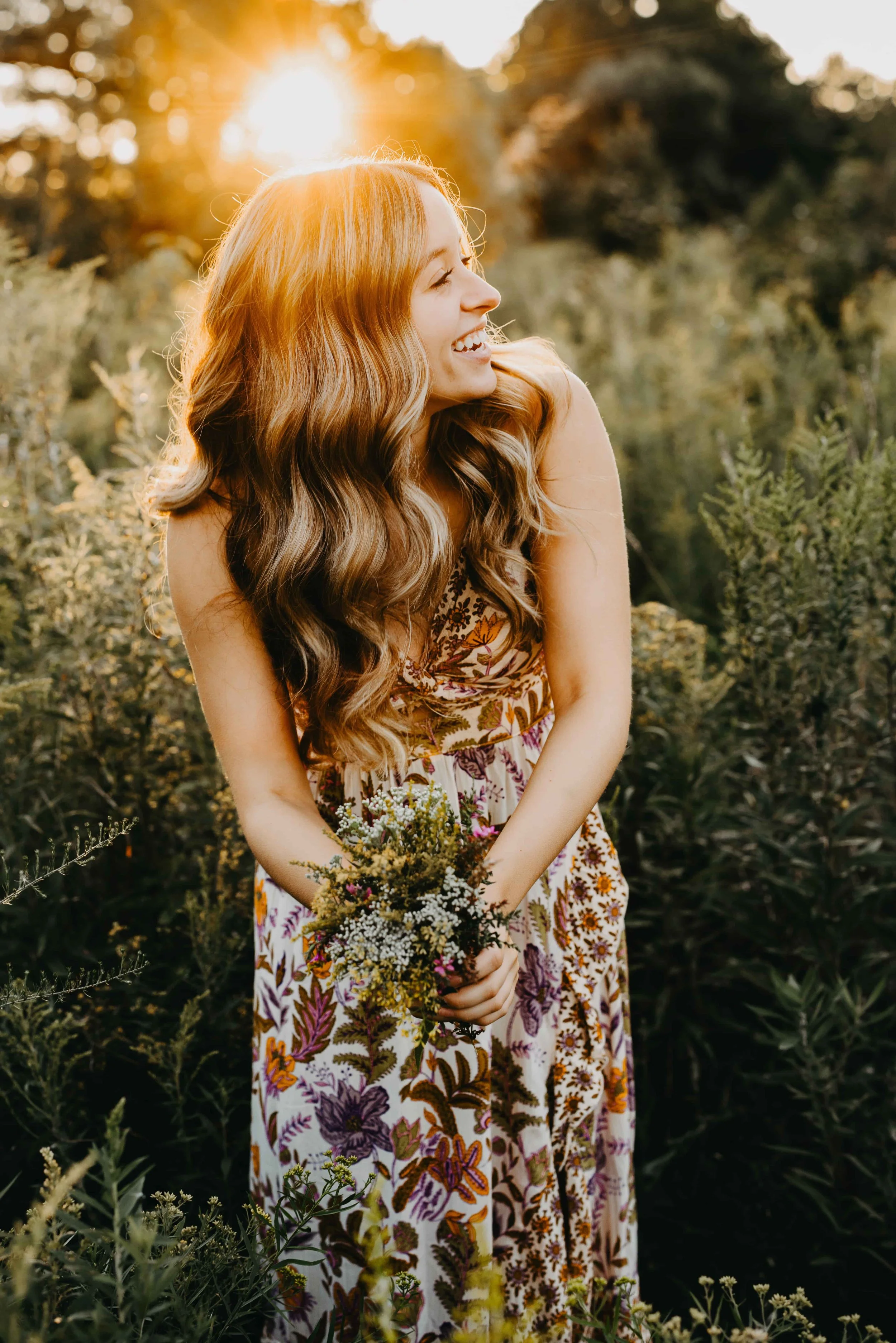 High school senior girl laughing and holding wildflowers in a field during golden hour in Cleveland, wearing a bohemian floral dress.