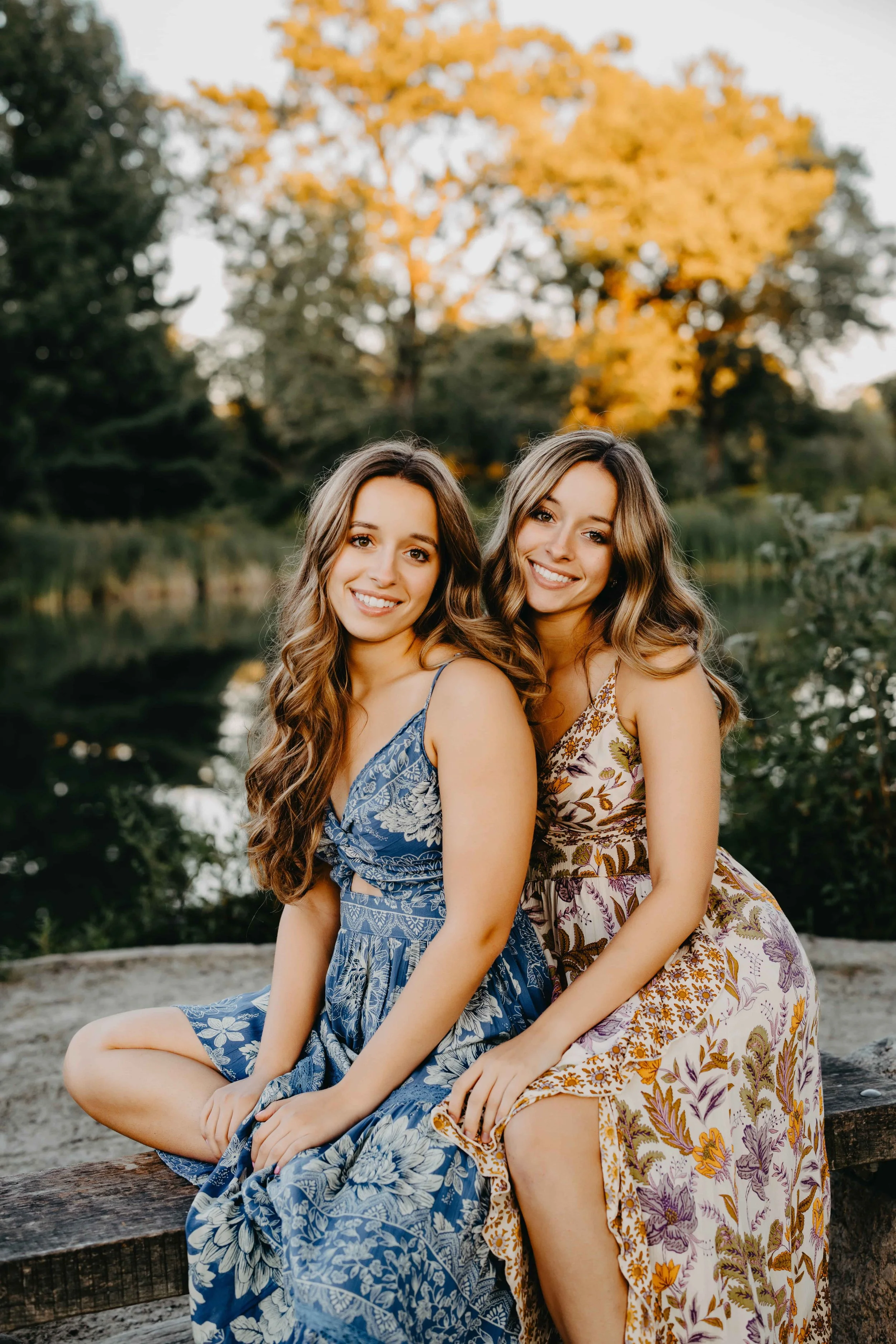 Twin sisters sitting together on a wooden bridge by a pond during fall in Cleveland, one in a blue floral dress and the other in a warm-toned floral dress.