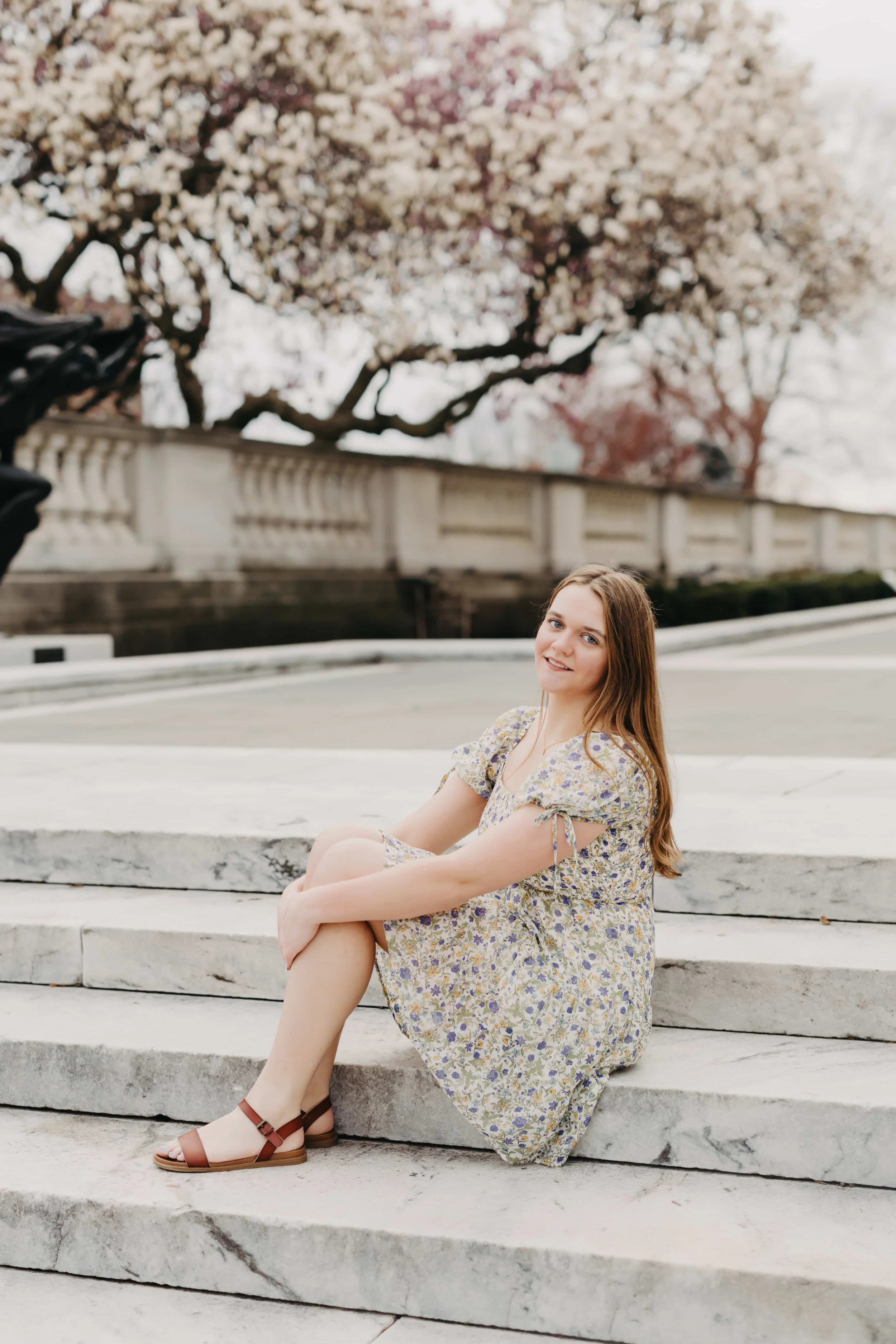 High school senior girl sitting on marble steps in front of a blooming magnolia tree at a Cleveland landmark, wearing a floral dress and brown sandals.