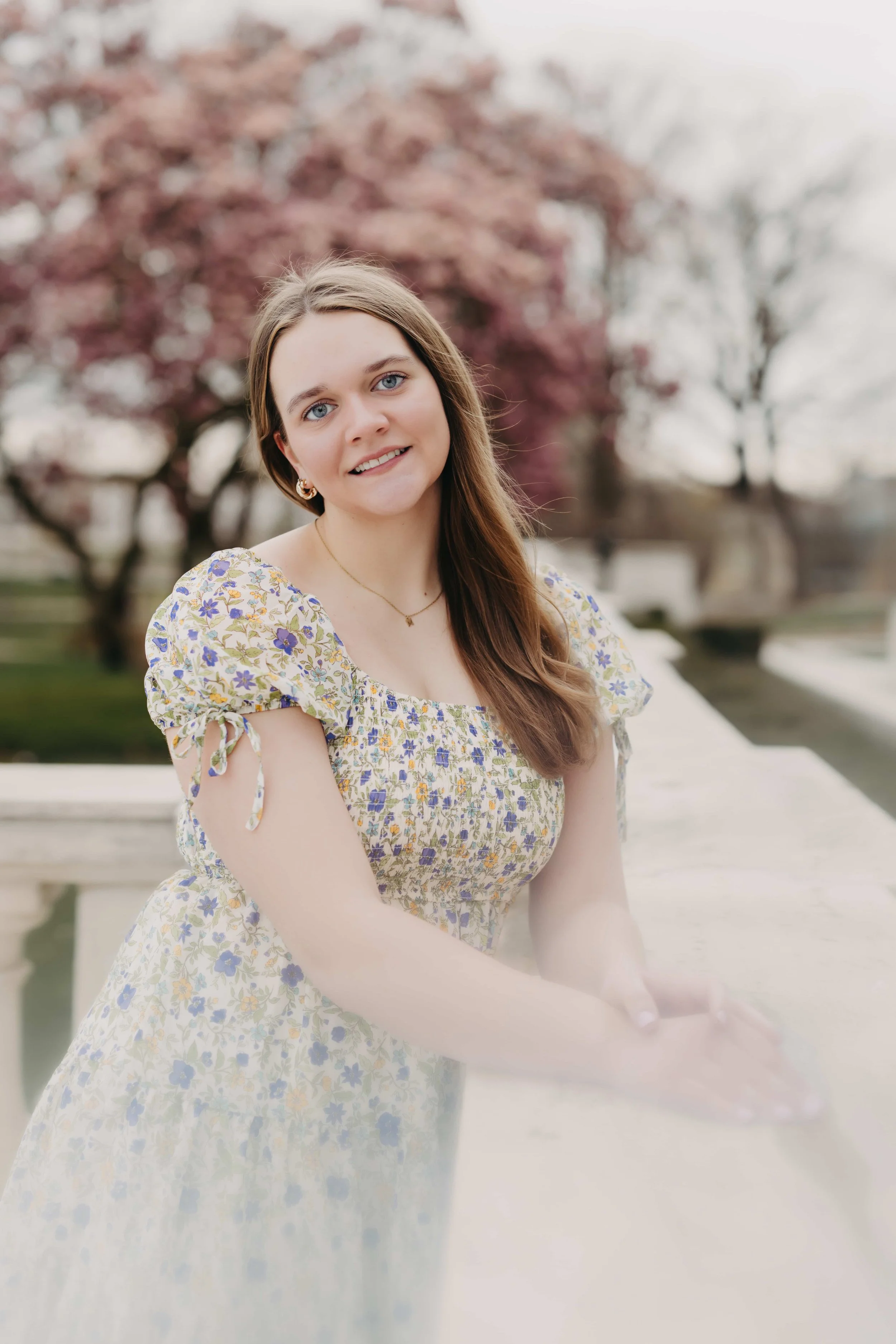 High school senior girl leaning against a white stone railing with pink cherry blossom trees in the background in Cleveland, Ohio, wearing a floral puff-sleeve dress.