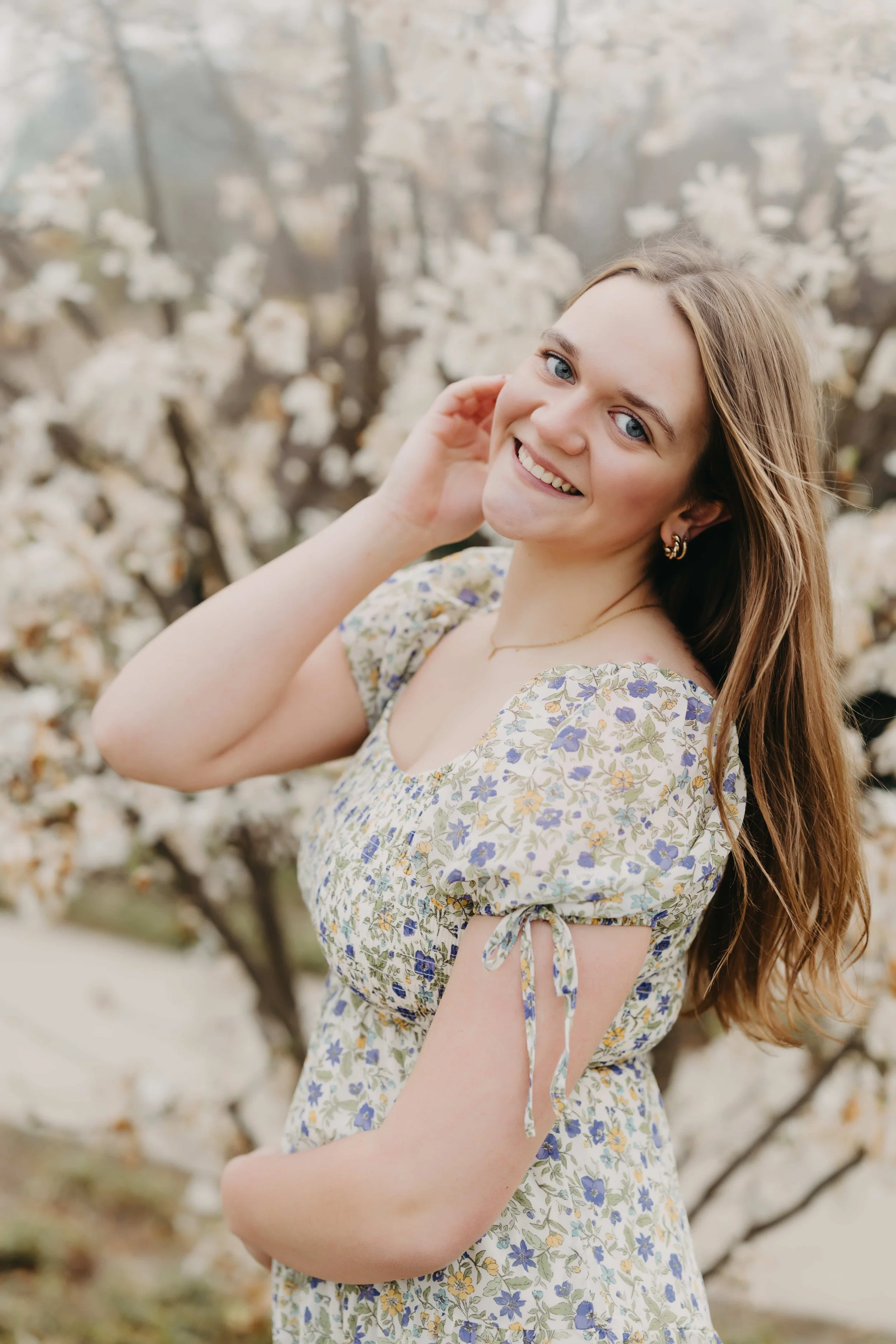 Close-up portrait of a high school senior girl smiling in front of white flowering trees in Cleveland, wearing a blue and green floral dress.