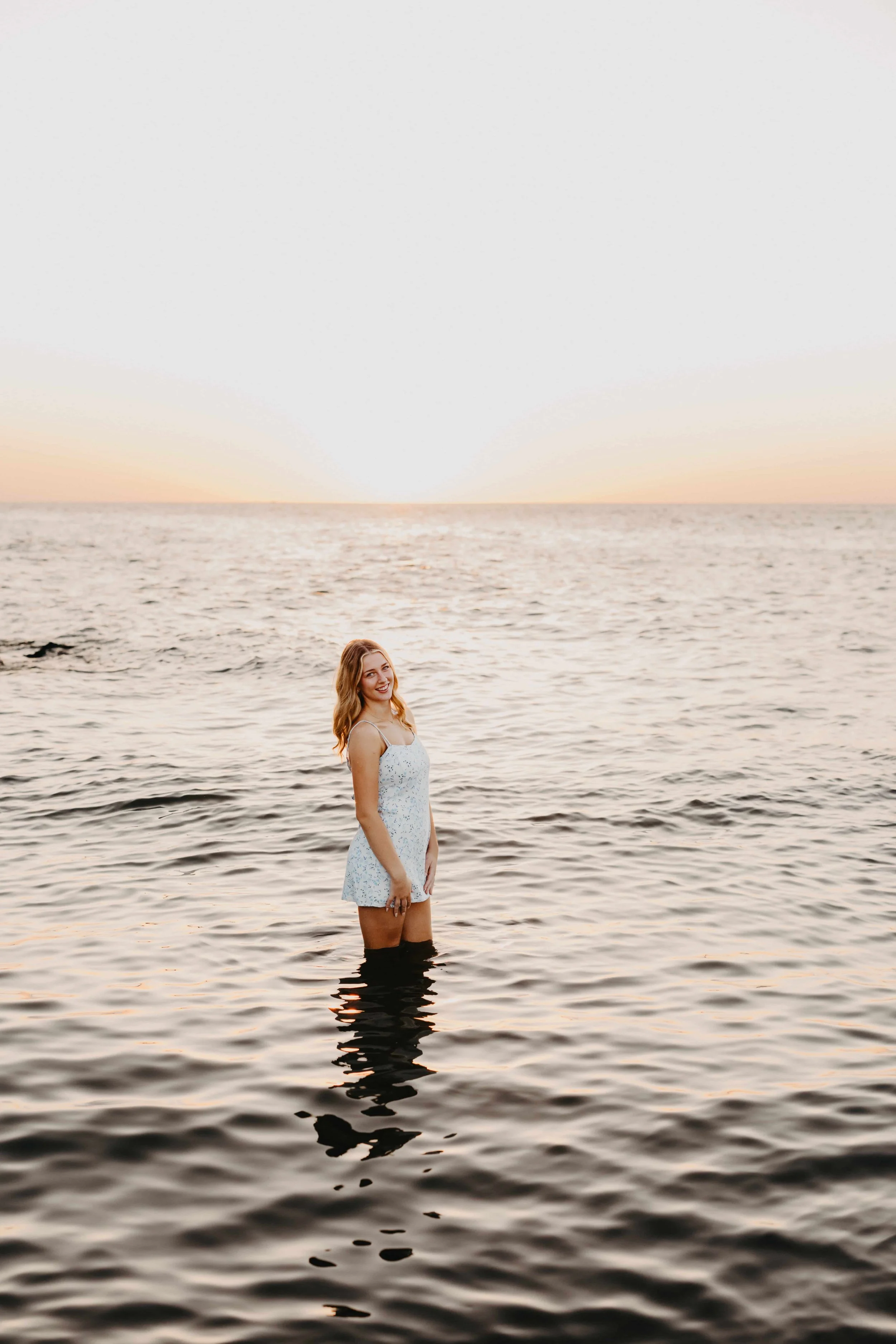 High school senior girl walking barefoot along a Cleveland beach at golden hour with large rocks and Lake Erie waves behind her, wearing a light blue floral mini dress.