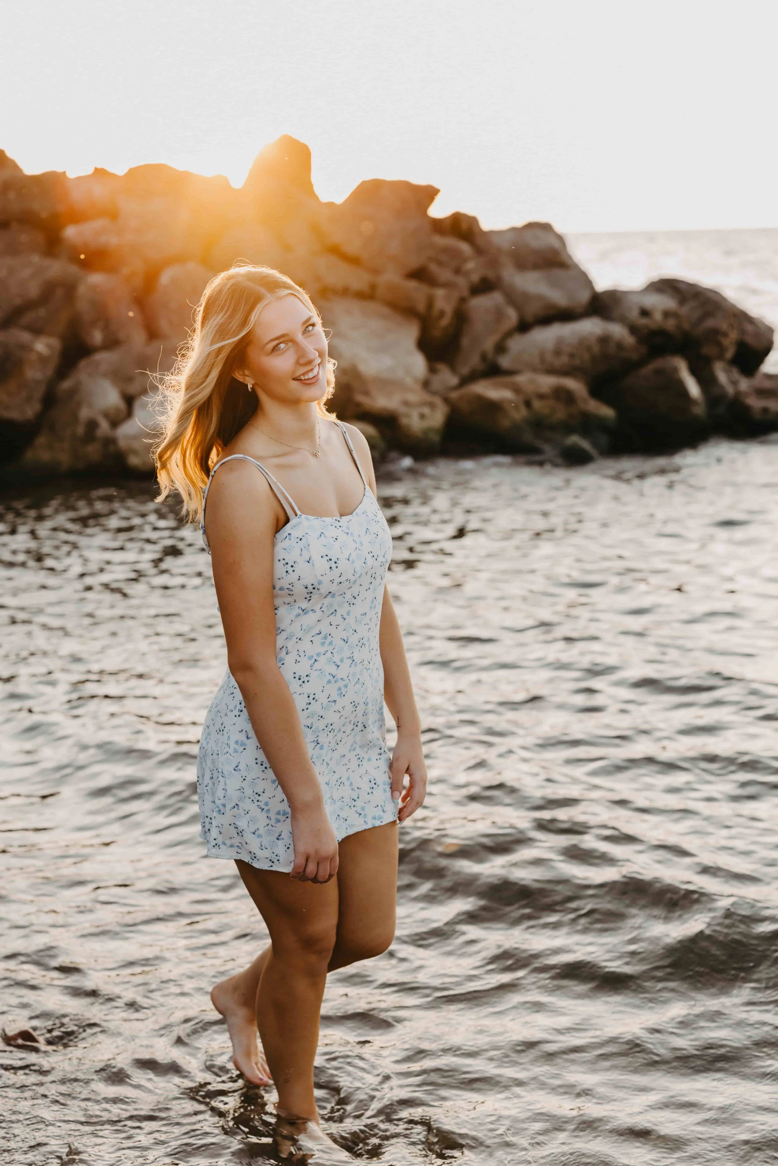 High school senior girl standing knee-deep in Lake Erie at sunset, wearing a light blue floral sundress with a pastel sky reflecting on the water.