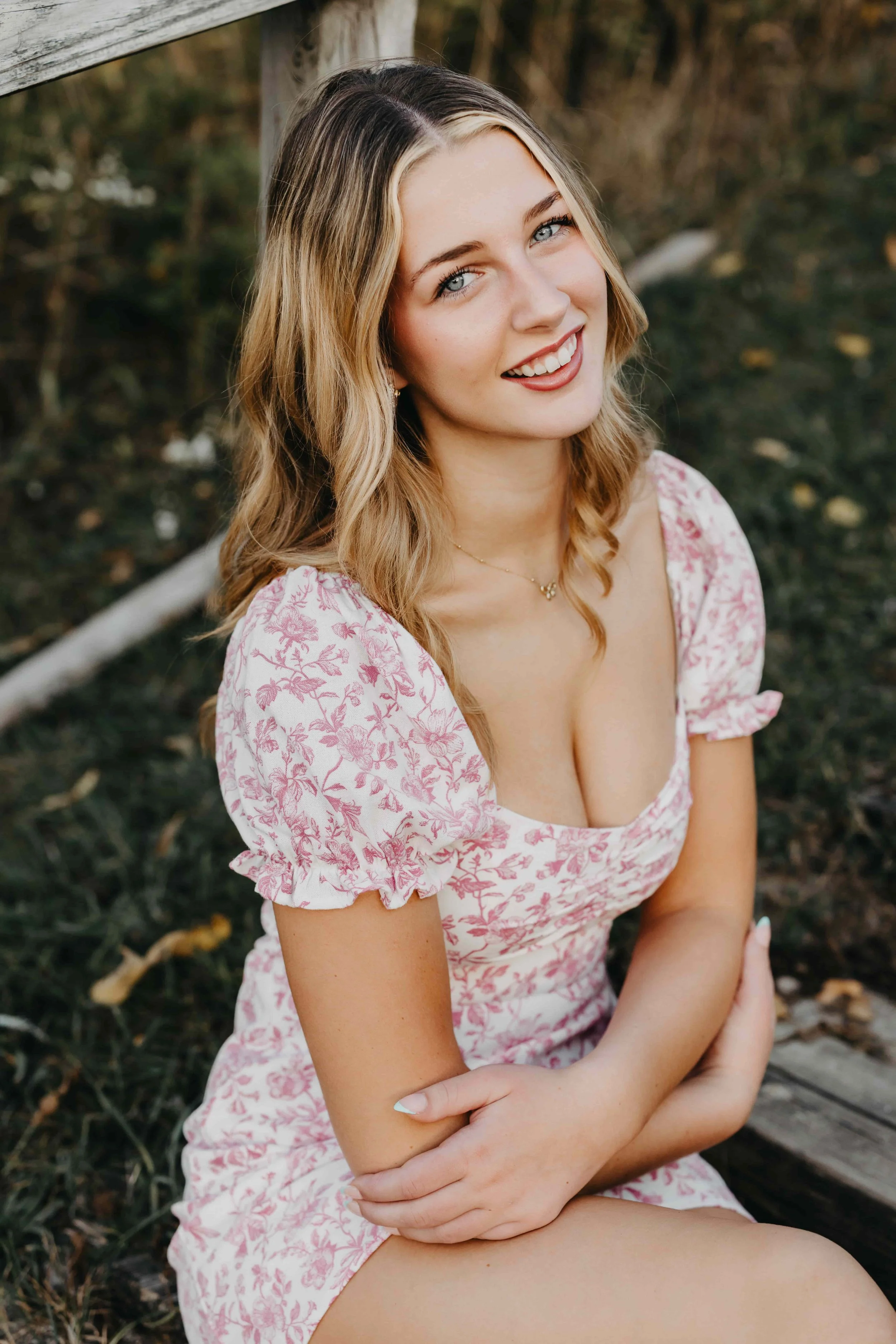 High school senior girl sitting on a rustic wooden bench in a natural outdoor setting in Cleveland, Ohio, wearing a white and pink floral puff-sleeve dress and smiling at the camera.