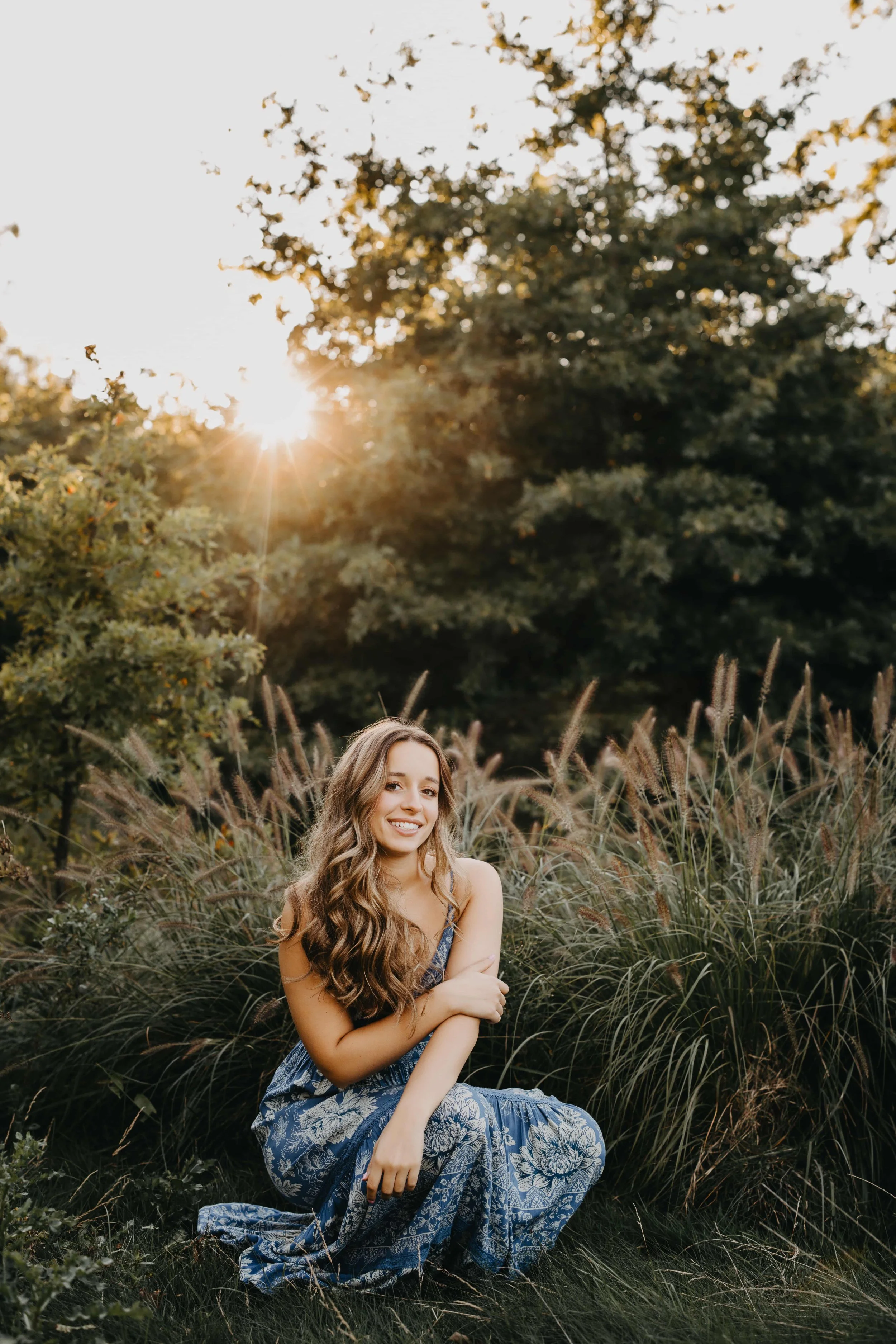 High school senior girl sitting in tall grass during sunset in Beachwood, Ohio, wearing a blue and white floral maxi dress.