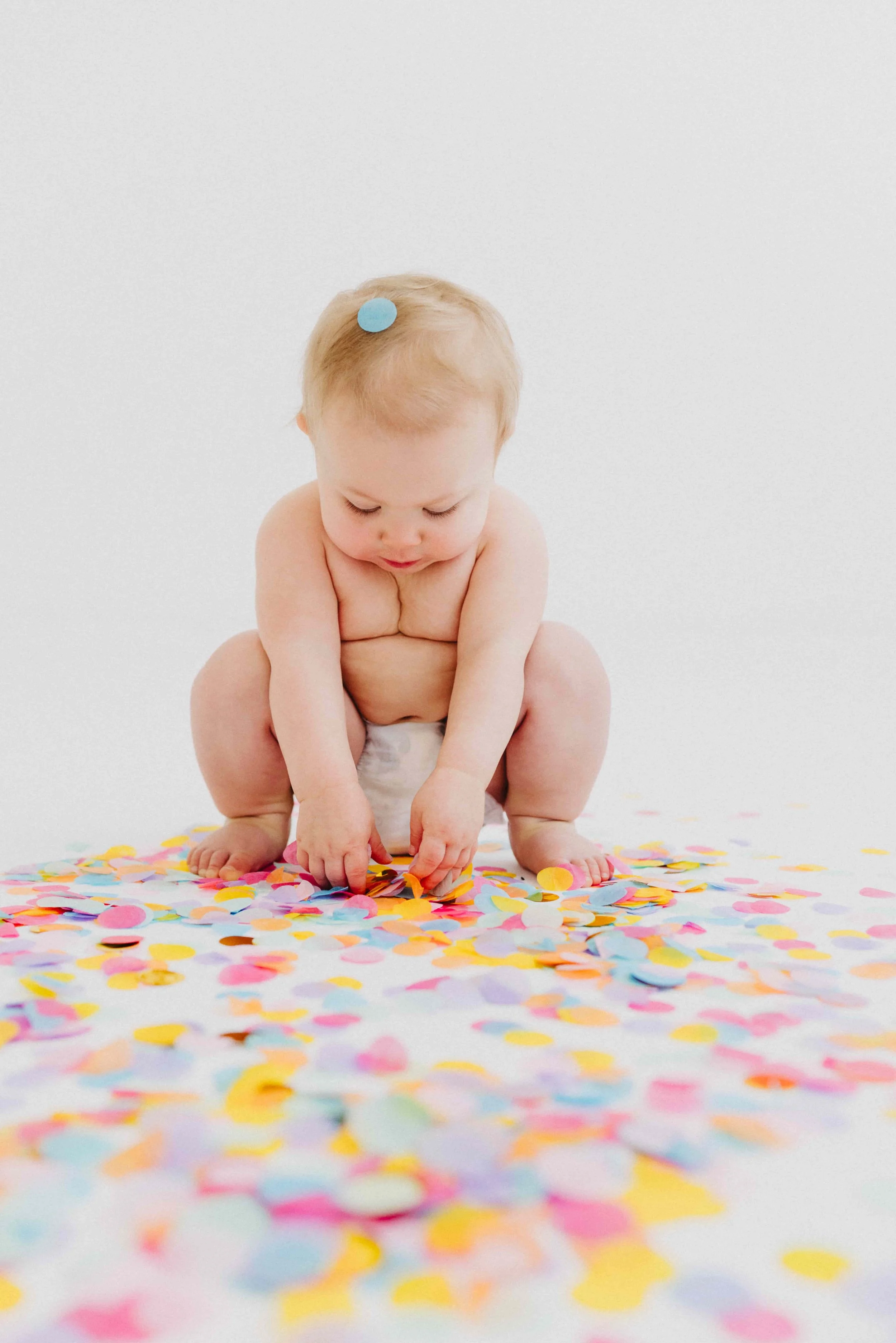 A toddler with light skin and blonde hair, wearing only a diaper, sits on the floor surrounded by colorful confetti and picks up some pieces.