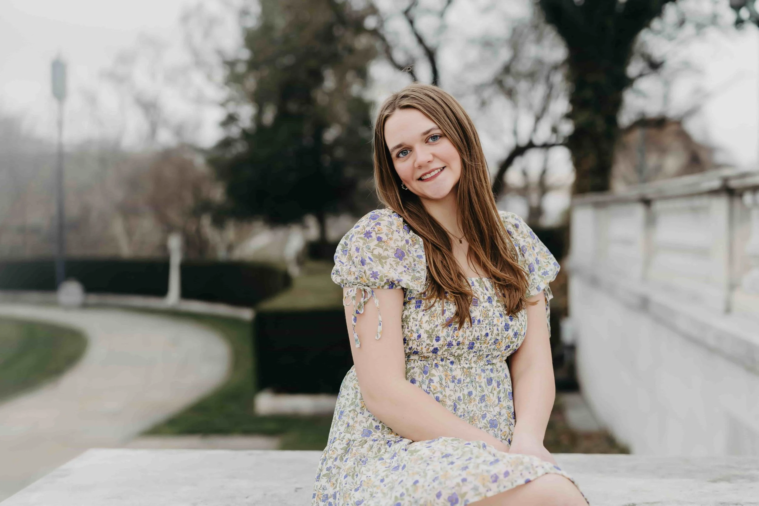 A young woman with long brown hair, wearing a floral dress, sitting outdoors near a sidewalk and a white fence, smiling at the camera.