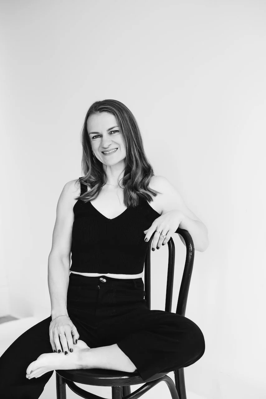 Black and white photo of a woman sitting cross-legged on a chair with a plain background. She is smiling, wearing a sleeveless black top and black pants.