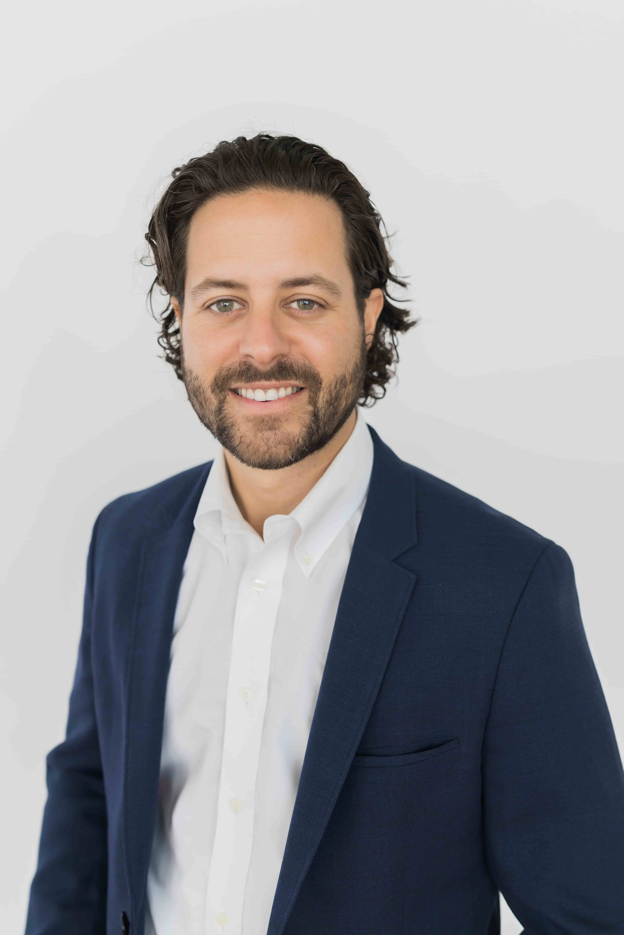 Headshot of a man with dark hair, beard, wearing a white shirt and navy blazer, smiling, against a plain light gray background.