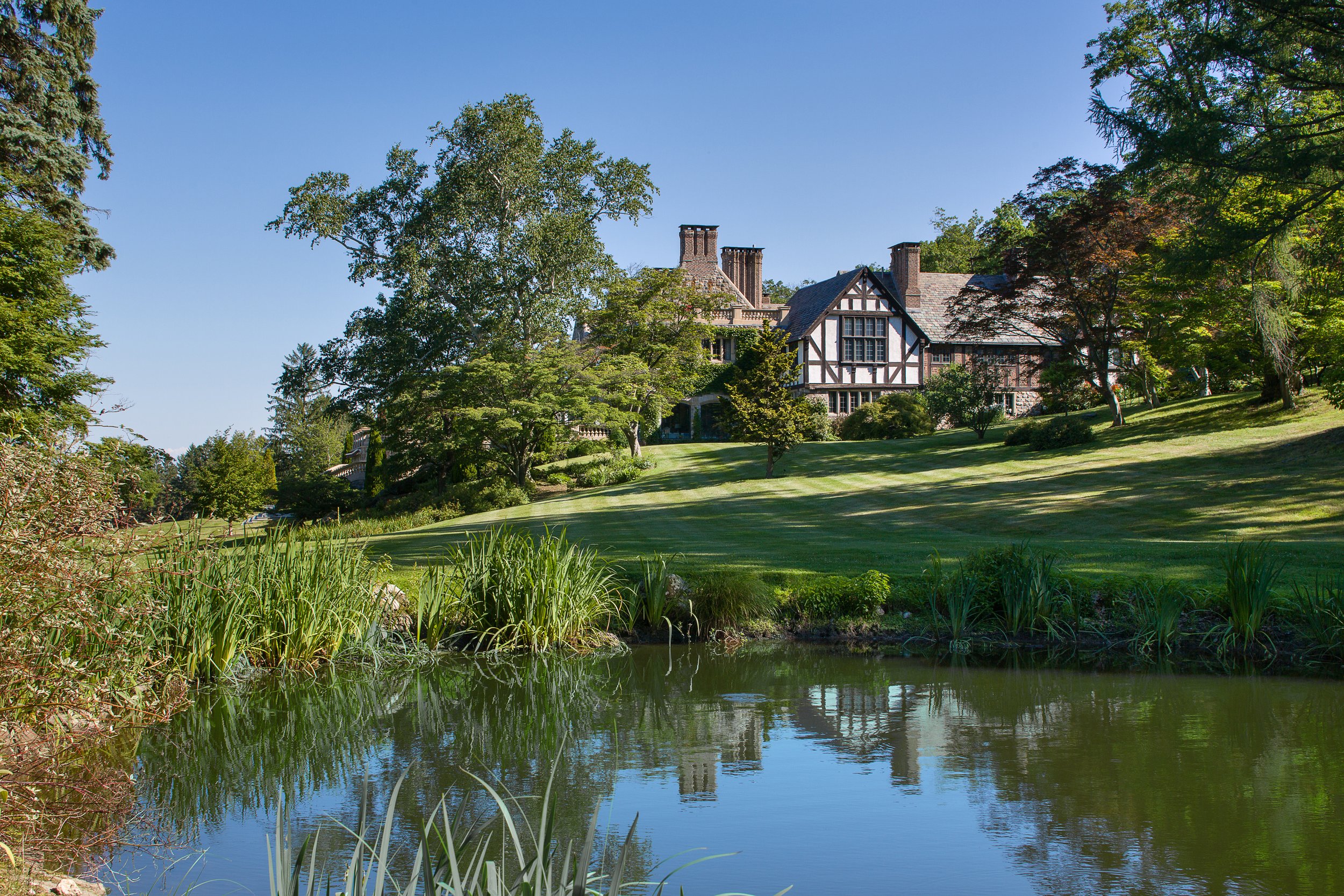 Large Tudor-style estate nestled on a hillside surrounded by lush greenery, overlooking a serene pond with reeds — Global Wealth Office showcasing timeless European architecture and luxury countryside living.