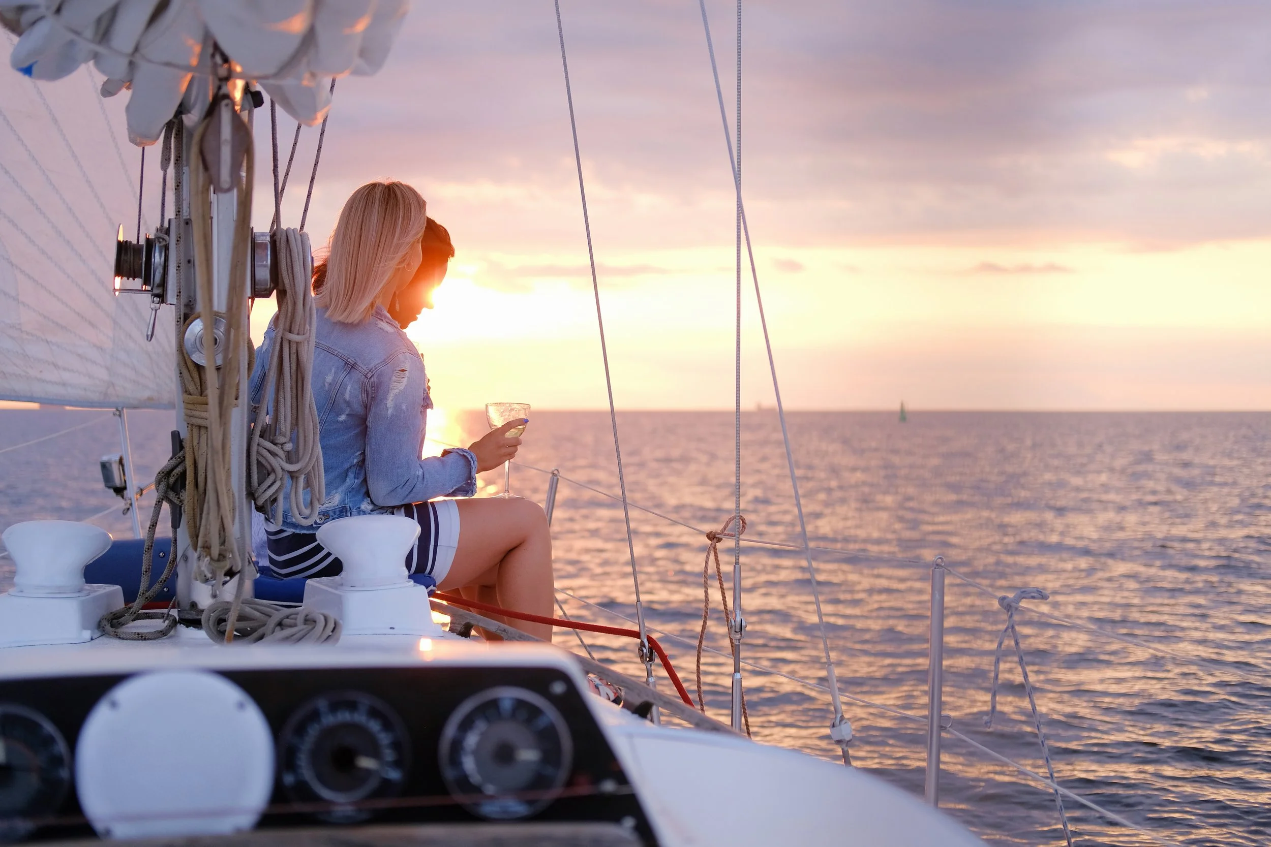 Two women sitting on a sailboat at sunset, one holding a glass of wine, with the ocean and a distant sailboat in the background.