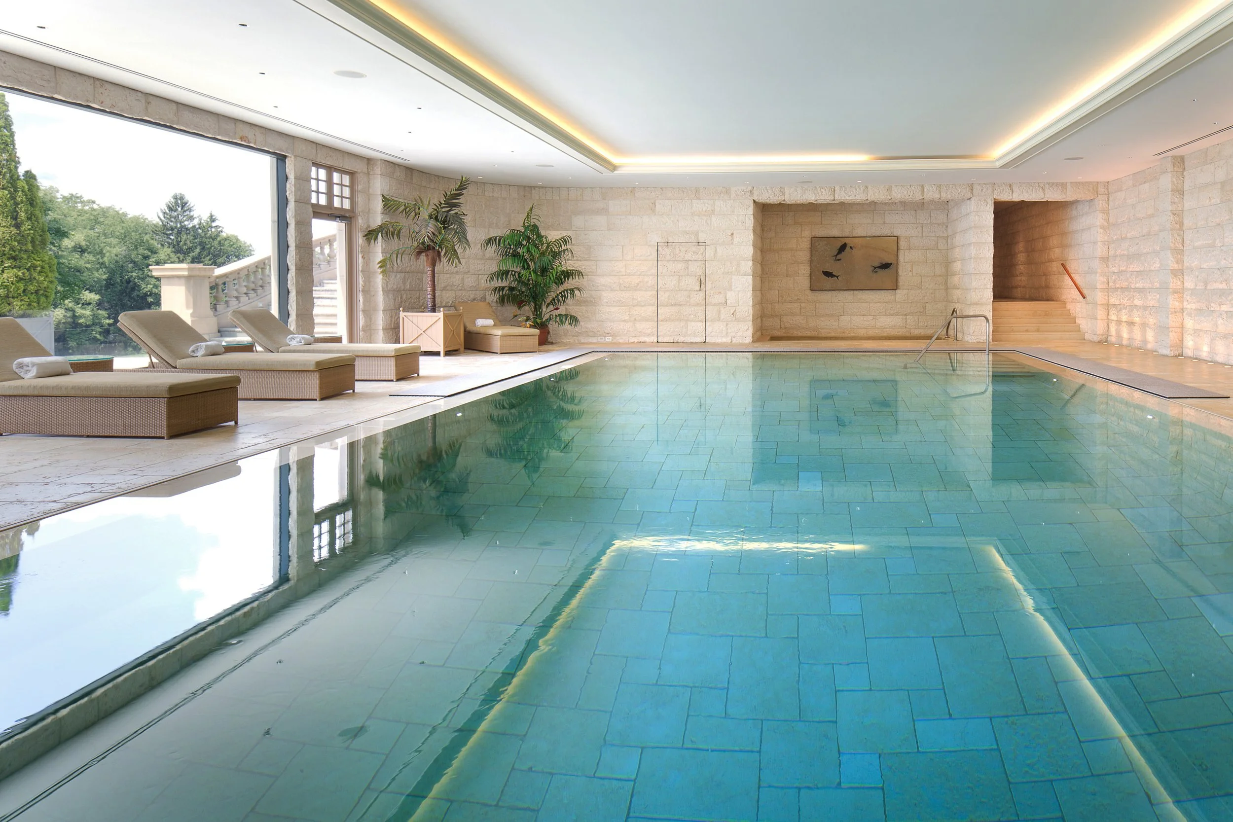 Indoor swimming pool surrounded by lounge chairs and lush plants, featuring large windows overlooking serene greenery — Global Wealth Interiors embodying spa-inspired tranquility and luxury wellness design.