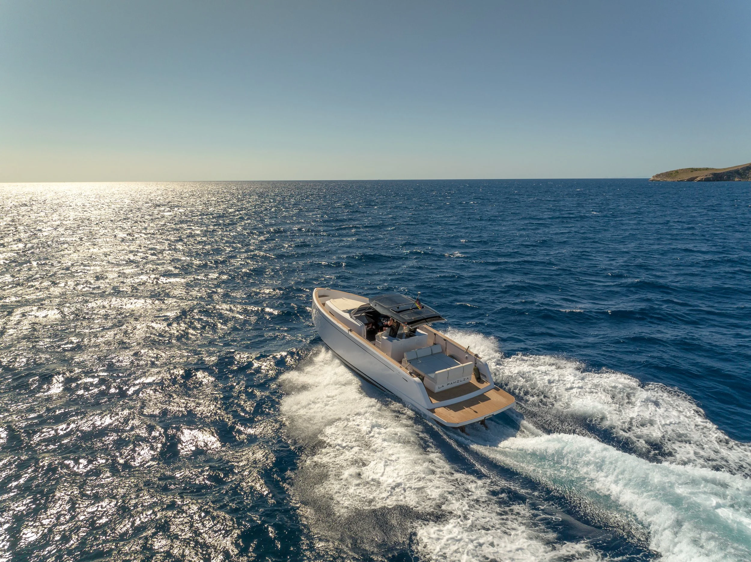 A motorboat speeding across the ocean with the shoreline and a hill in the distance under a clear blue sky.