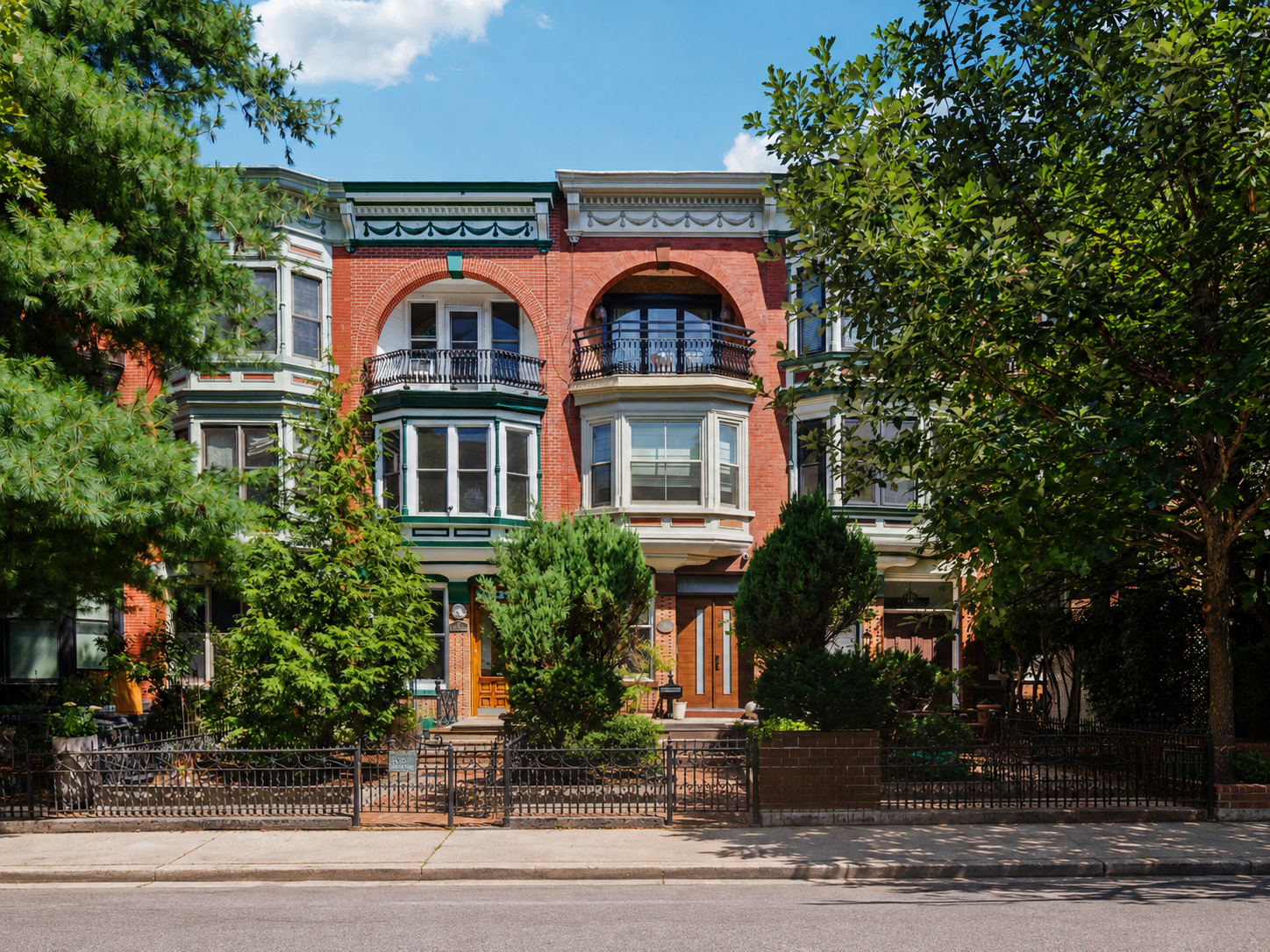 A row of historic Victorian-style brick houses with ornate trim, bay windows, and small front yards enclosed by black metal fences, set along a sidewalk with large leafy trees.