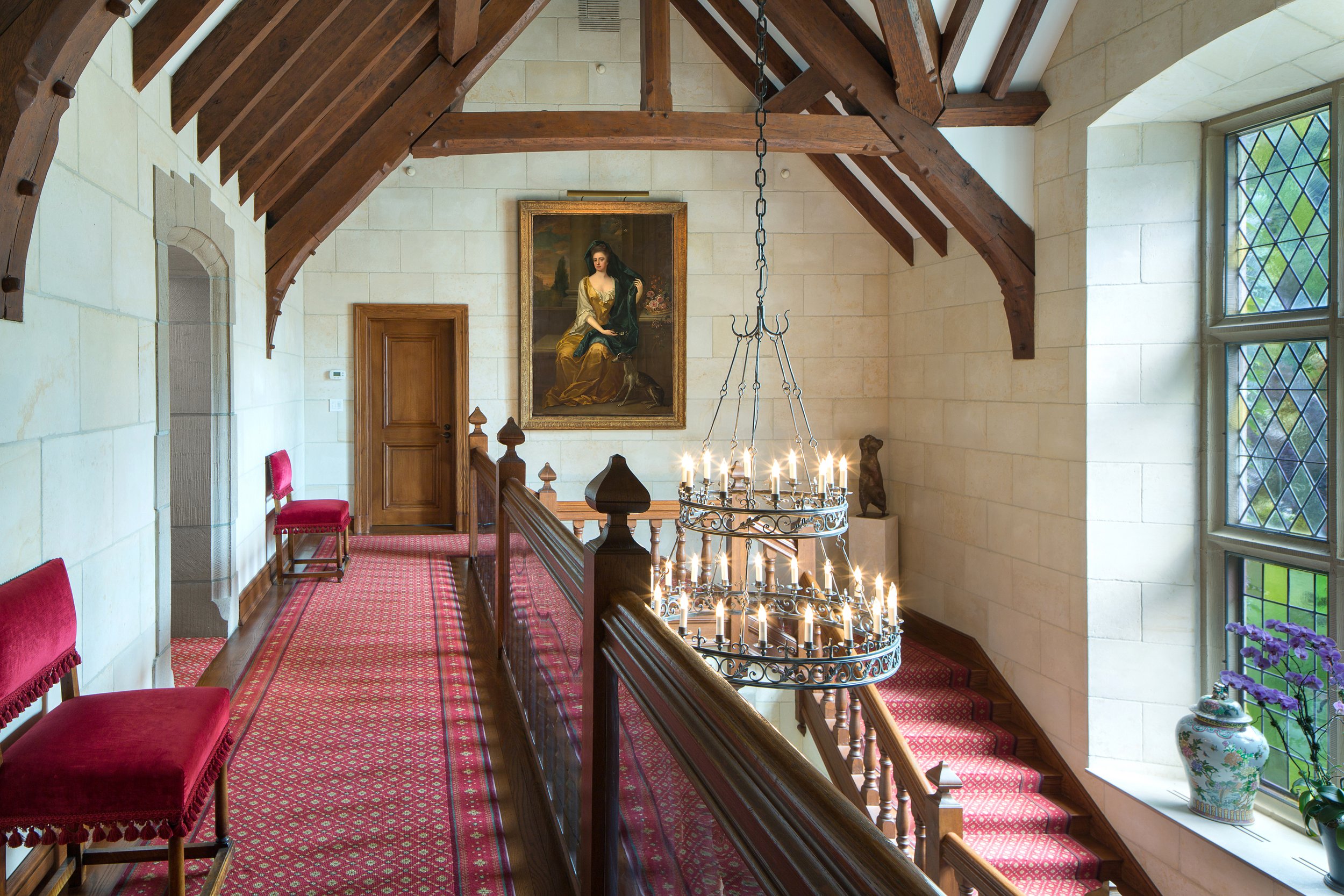 Grand staircase hall with chandelier, exposed wooden beams, stained glass windows, red carpet, and portrait painting — Global Wealth Interiors highlighting architectural grandeur and classic interior elegance.
