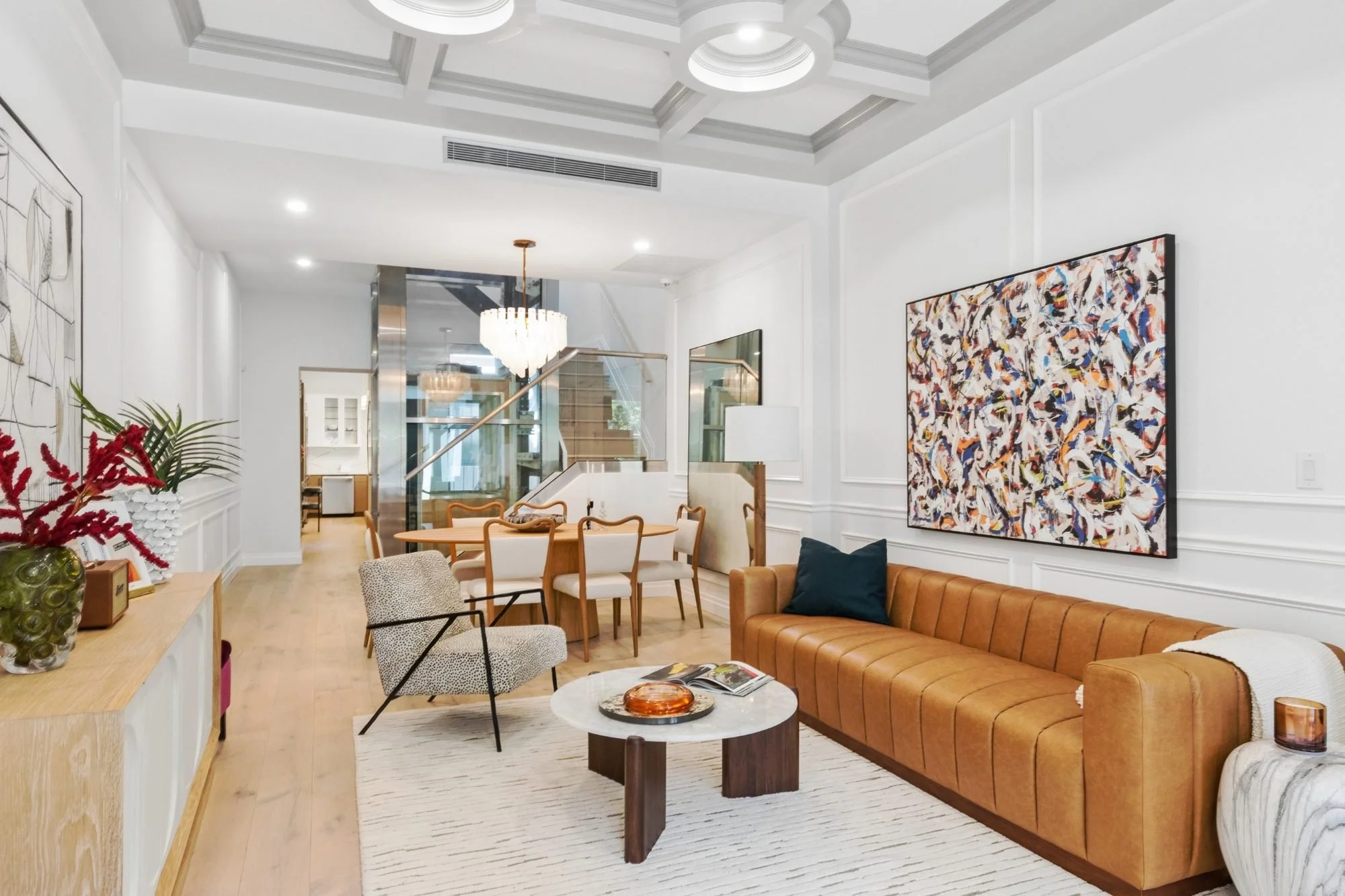 Living room of 143 Milton Street with white walls, wooden flooring, a tan leather sofa, two patterned armchairs, a white round coffee table with a decorative tray, artwork on the walls, a hanging chandelier, and a staircase leading to an upper floor.