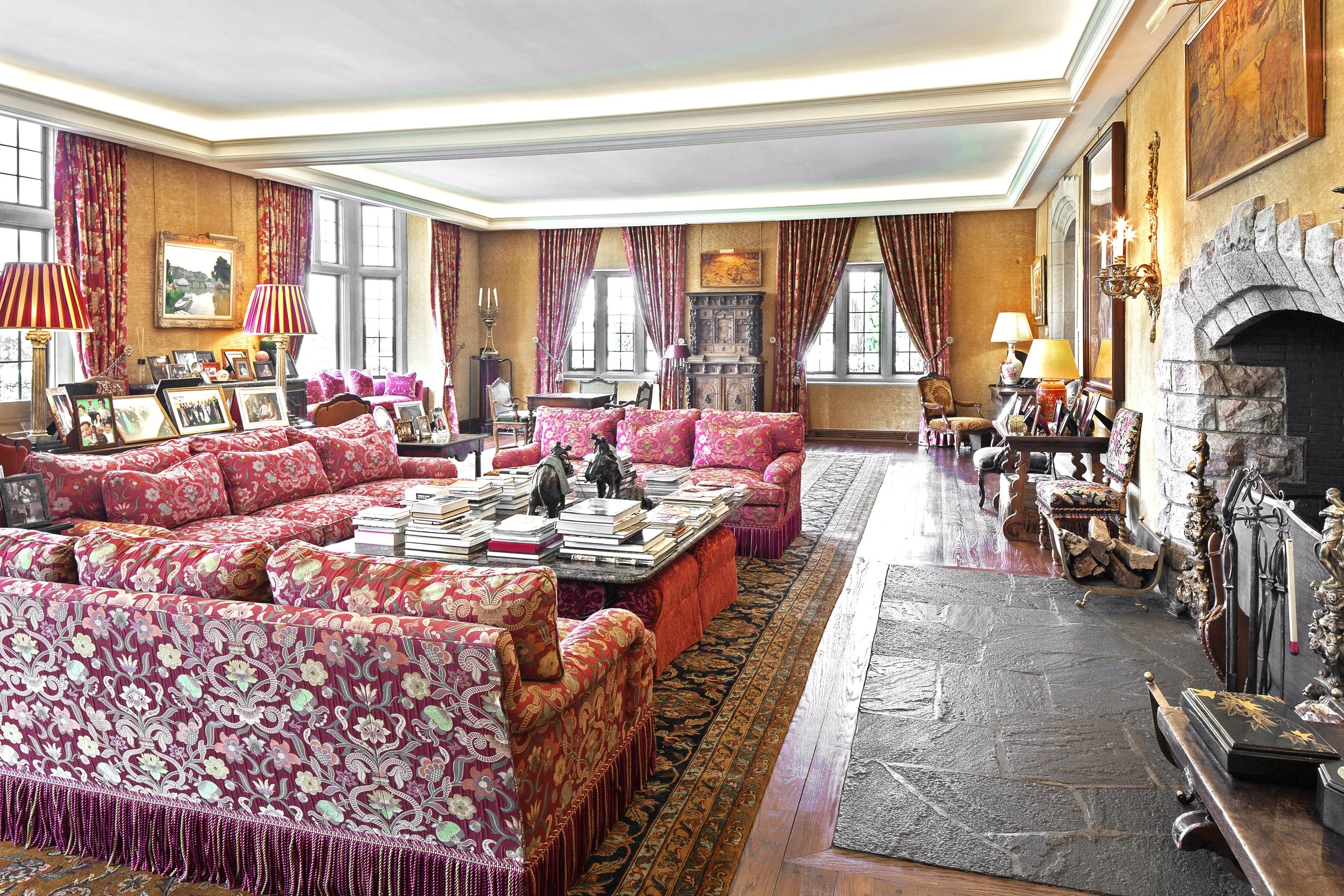 Spacious, elegant living room with floral upholstered sofas, stone fireplace, antique furniture, and red-and-gold draped windows adorned with framed photographs — Global Wealth Interiors showcasing timeless charm and curated classic design.