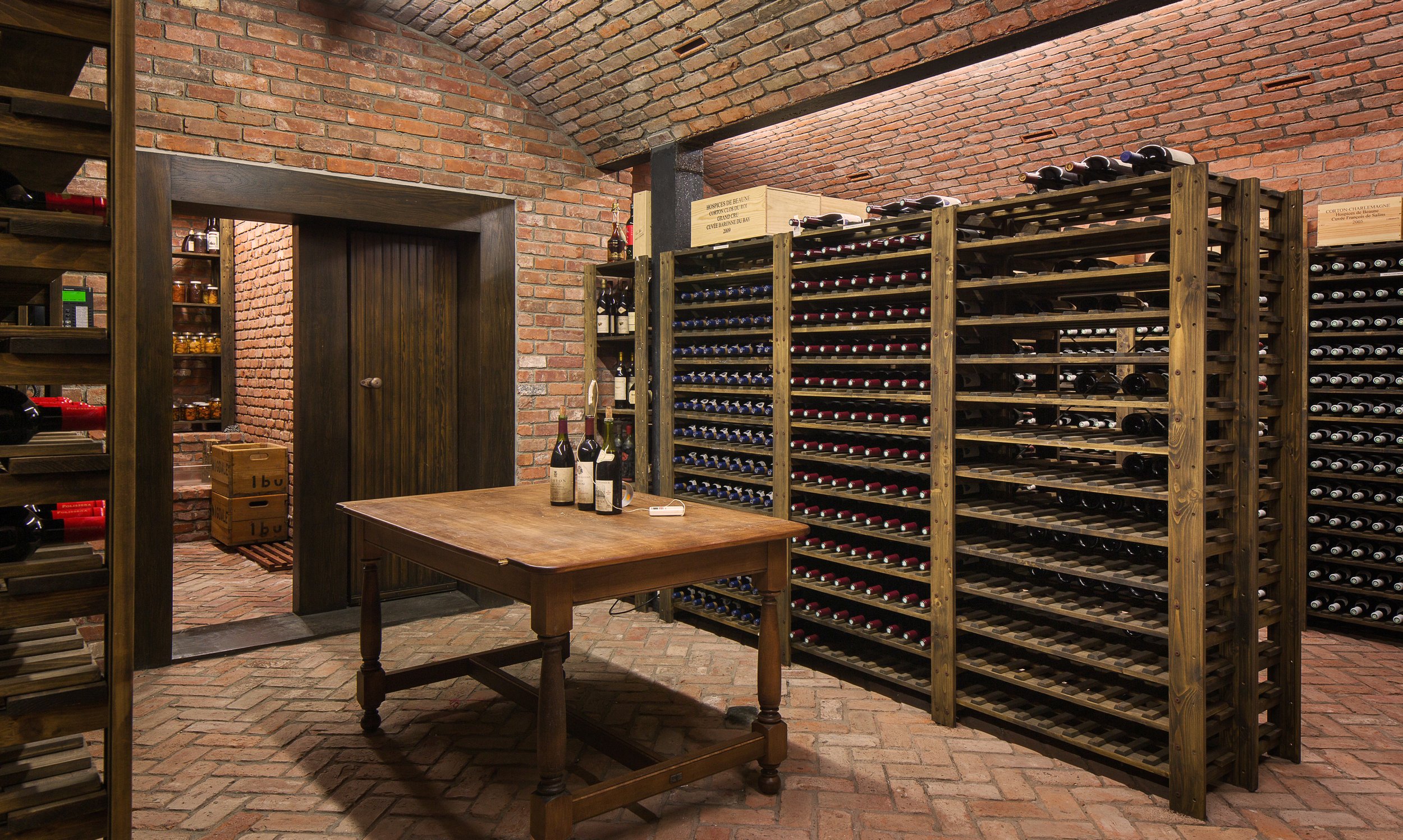 Wine cellar with brick walls and flooring, wooden racks filled with bottles, and a central wooden table displaying wine bottles — Global Wealth Interiors showcasing refined craftsmanship and timeless cellar design.