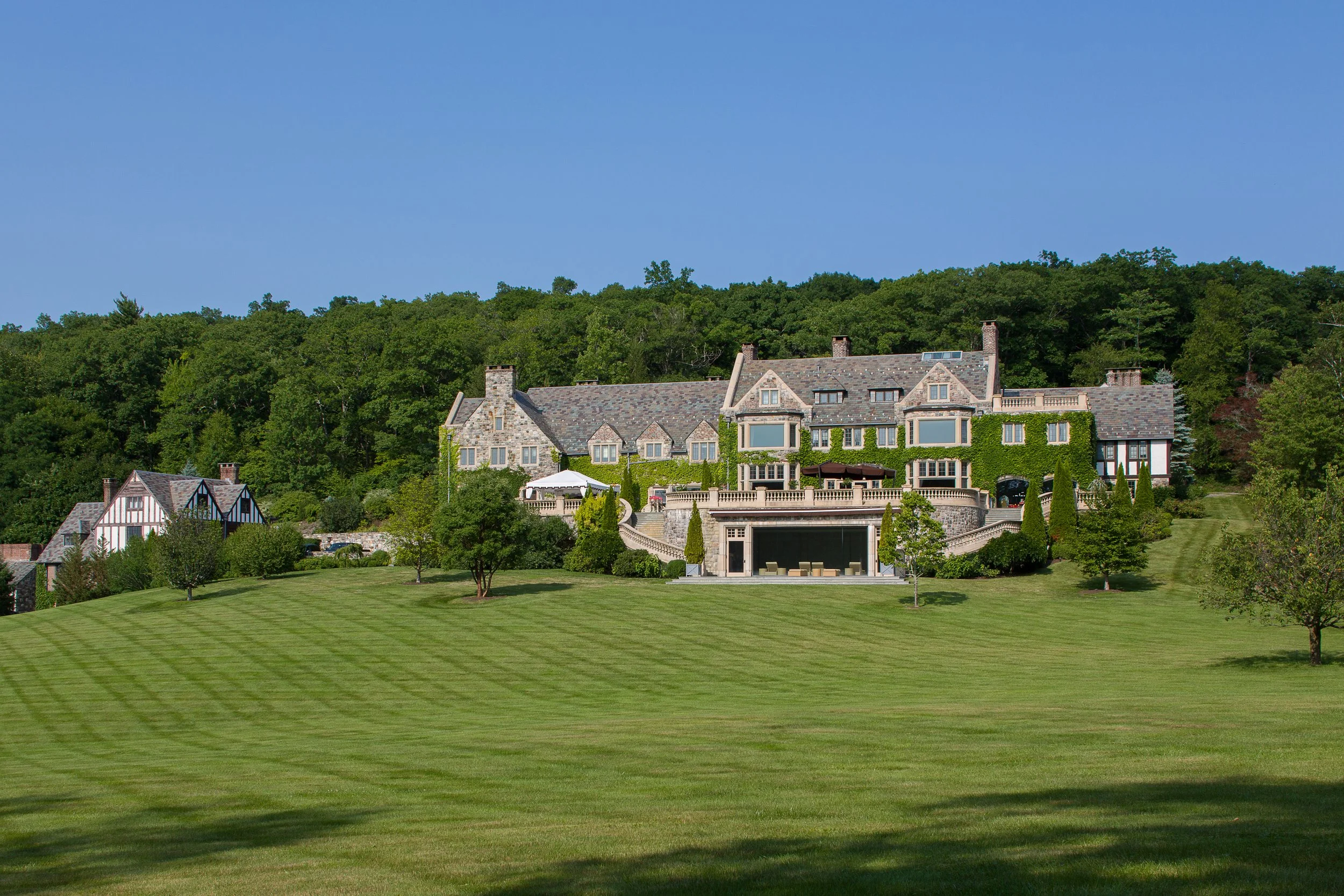 Grand mansion-style estate on a hill surrounded by manicured lawns and trees, with a forested backdrop under a clear blue sky — Global Wealth Office showcasing distinguished architecture and luxury countryside living.