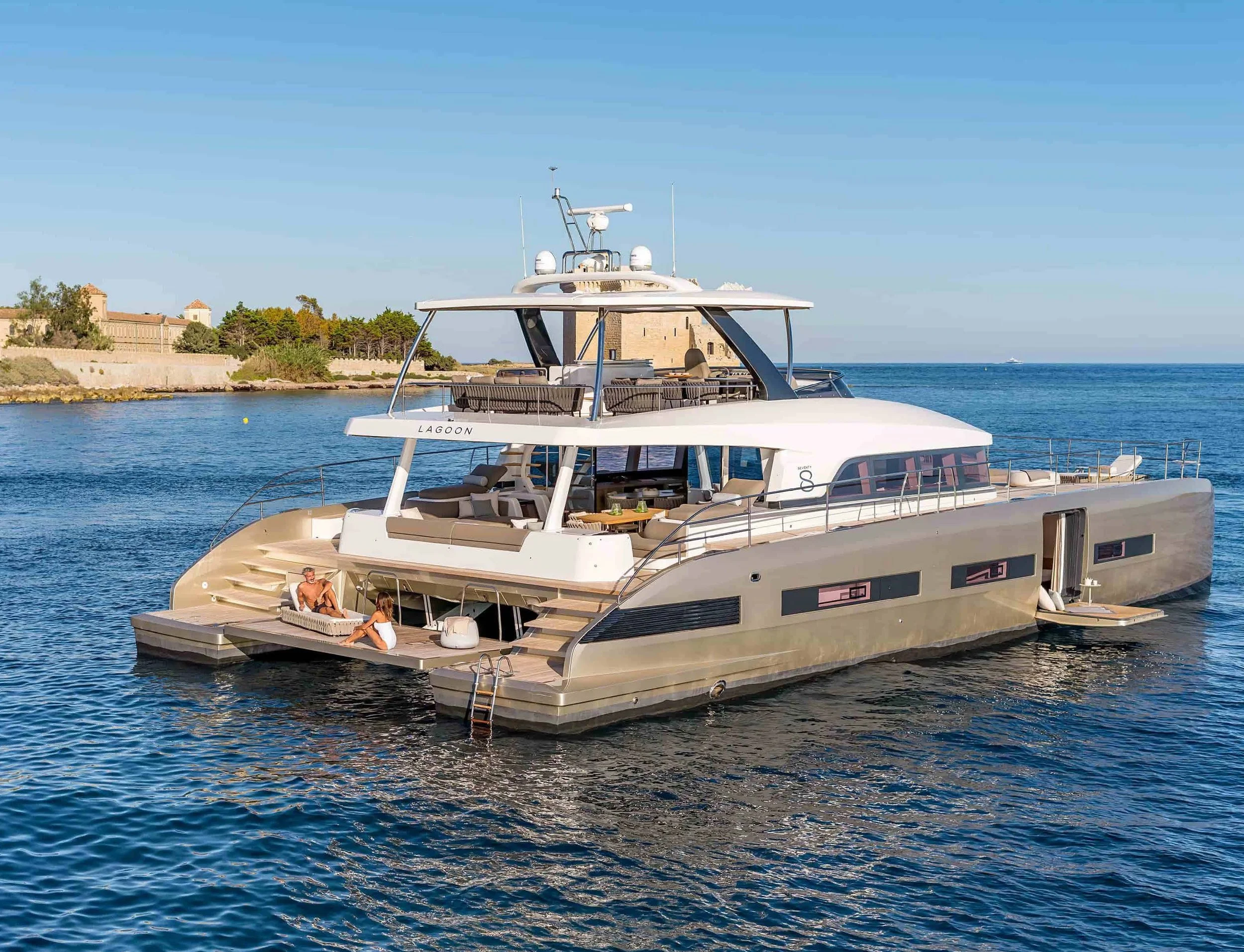 Global Wealth Maritima luxury yacht named Lagoon docked in calm blue water near a shoreline with historic buildings and lush green trees under a clear sky.